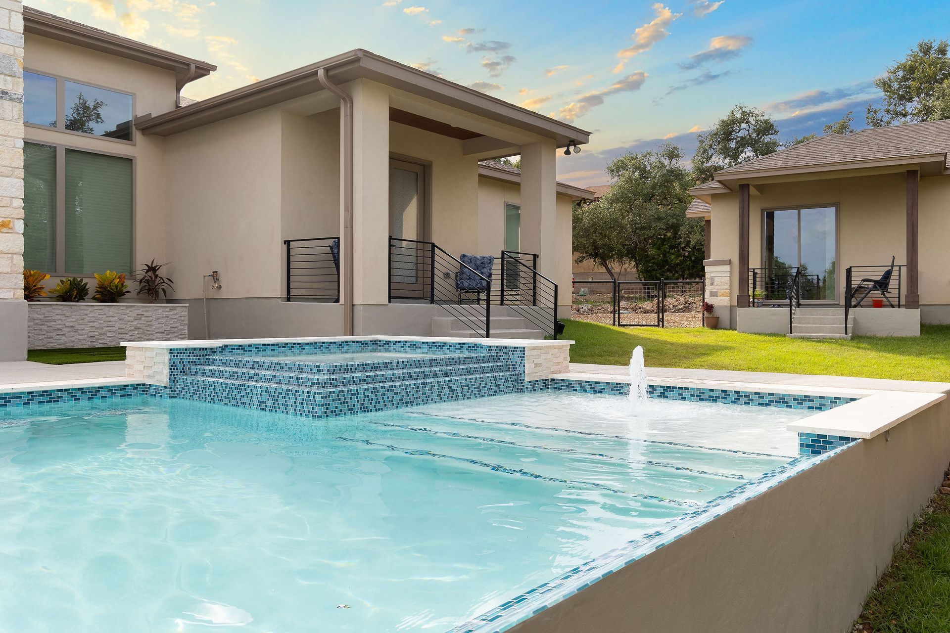 Modern backyard pool with spa, light stucco homes, and a small fountain at sunset