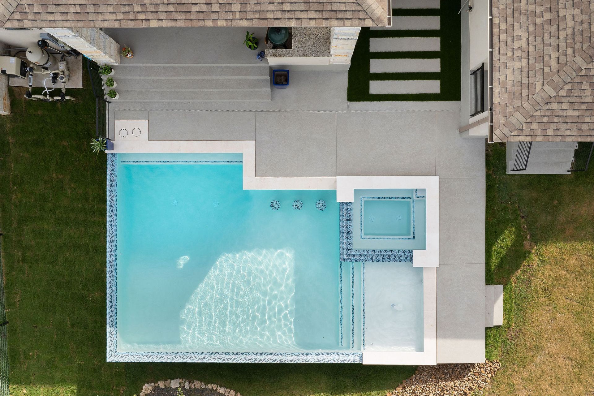 Aerial view of a modern backyard with a turquoise pool, spa, and white patio beside a house.