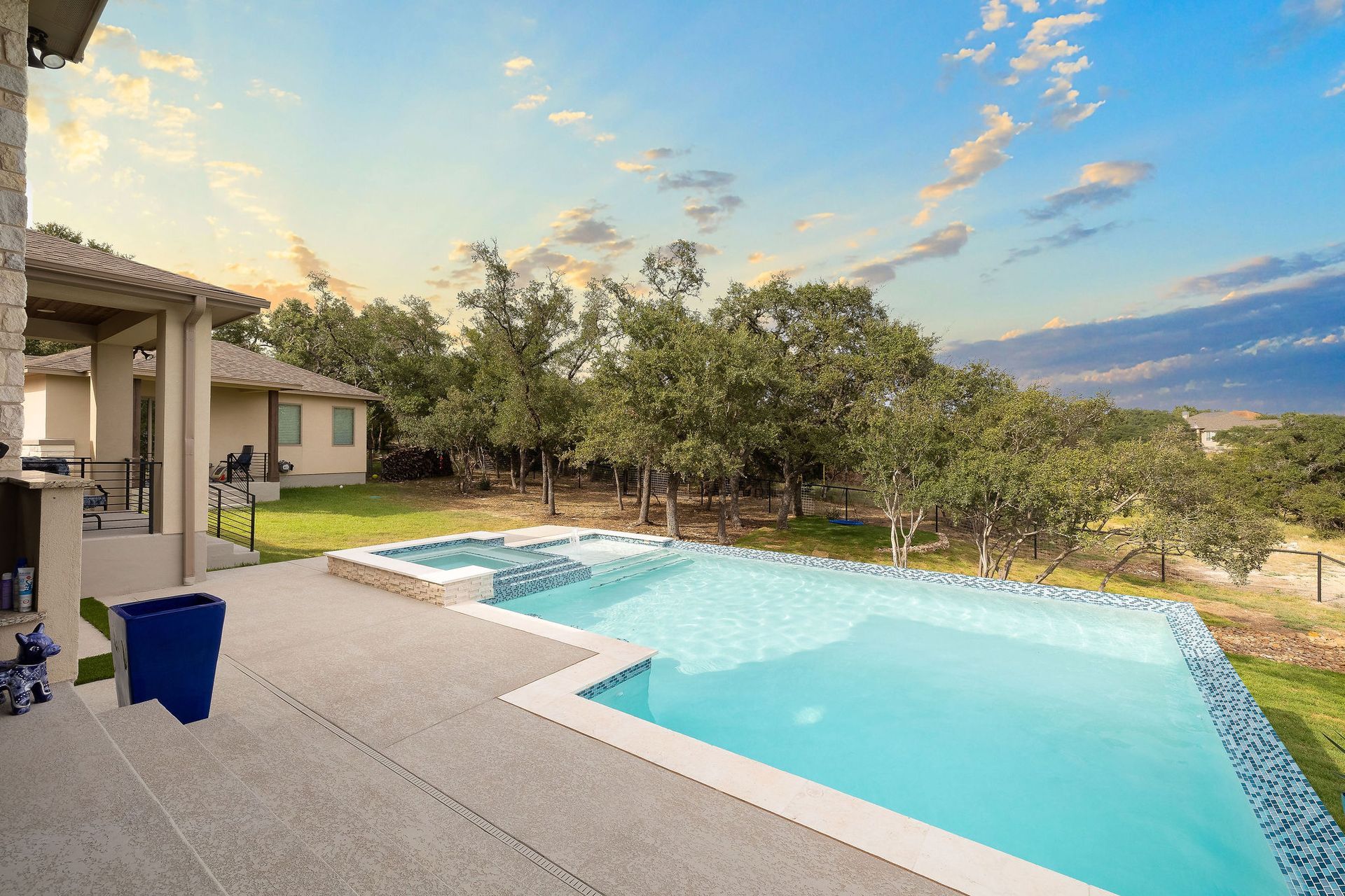Patio beside a turquoise swimming pool at sunset, with trees and a cloudy sky beyond.
