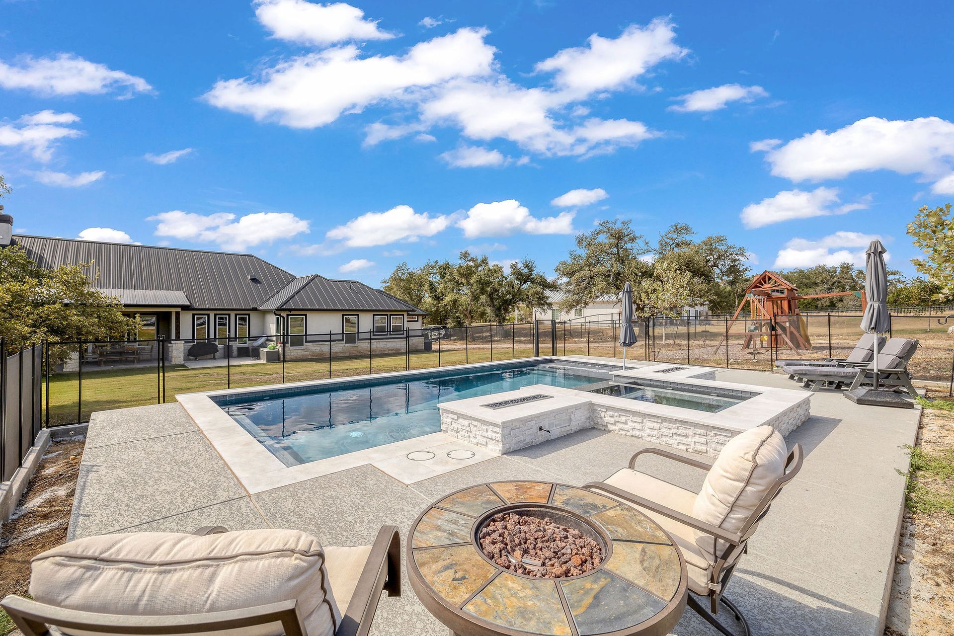Backyard patio with a rectangular swimming pool, lounge chairs, fire pit, and sunny blue sky.