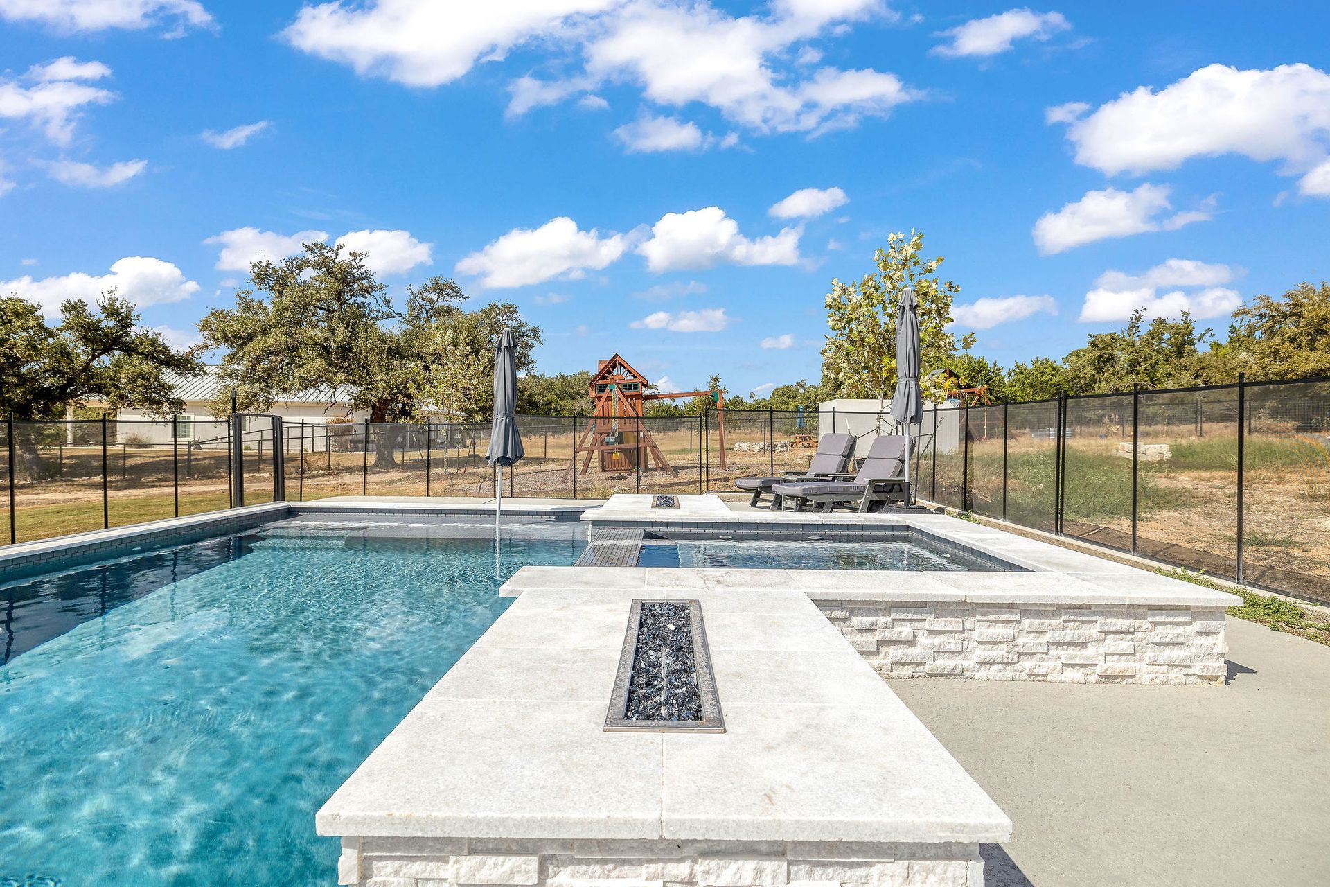Modern backyard pool with stone deck, lounge chairs, and fenced yard under a blue sky