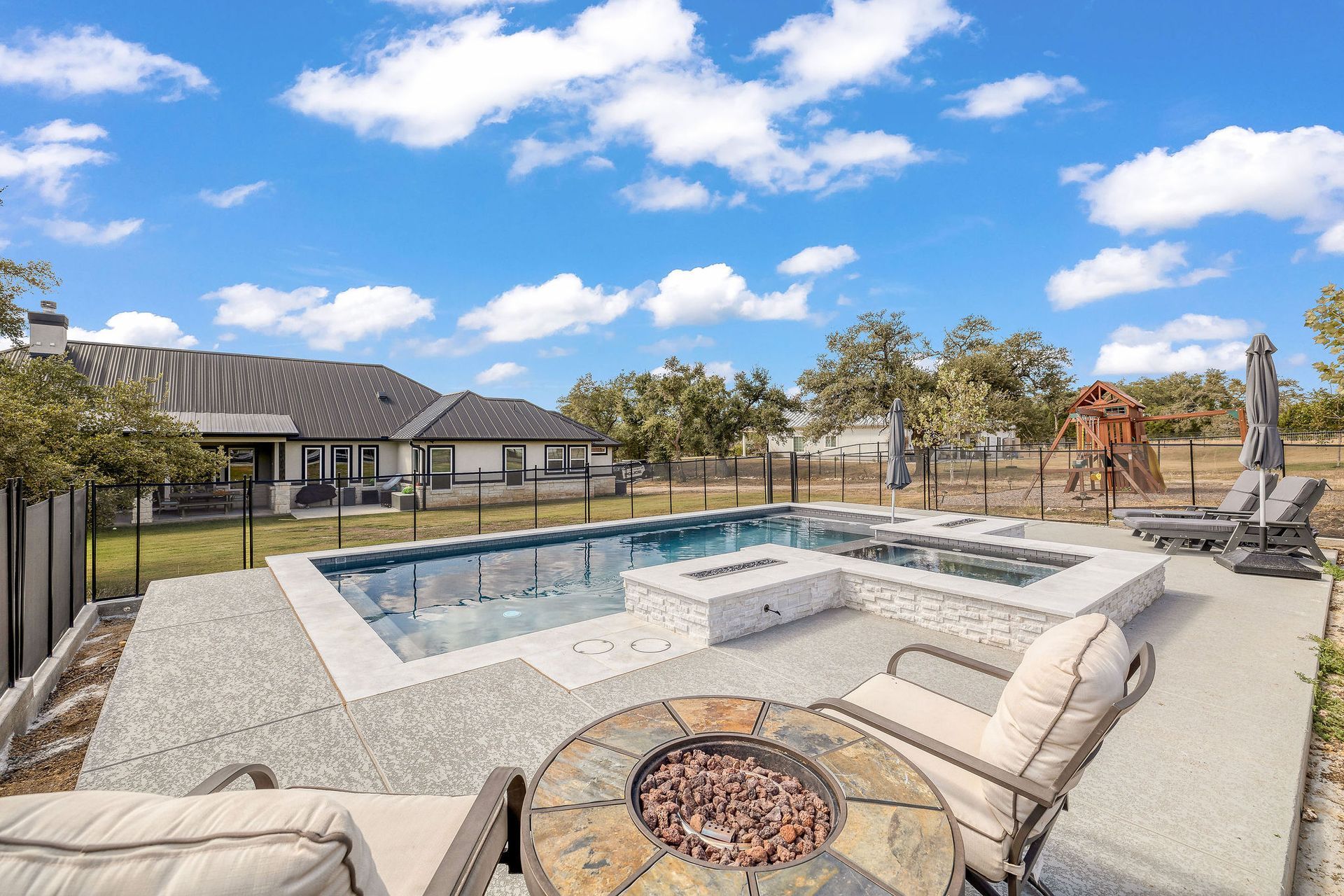 Backyard with a rectangular pool, stone patio, lounge chairs, fire pit, and blue sky with clouds