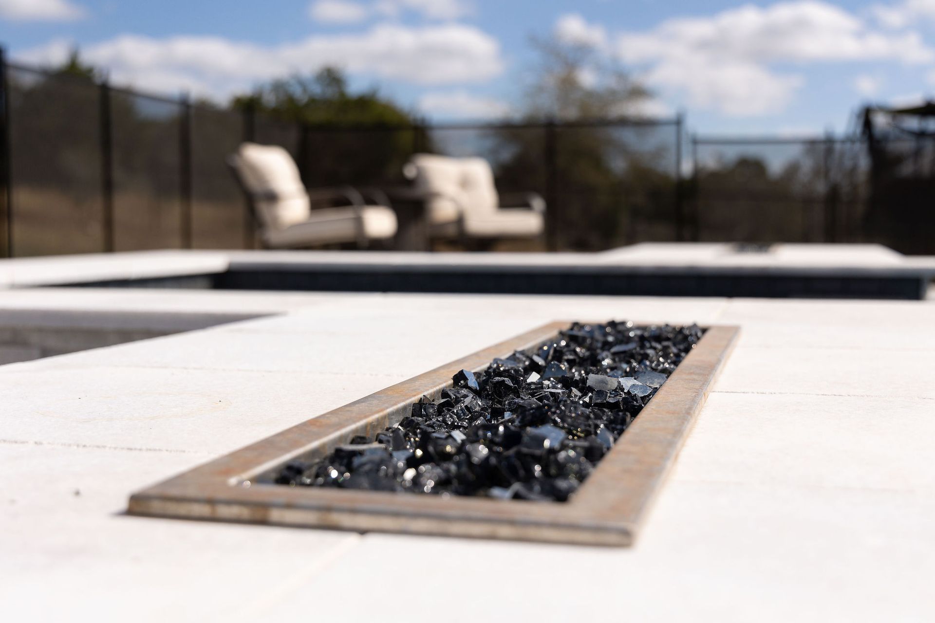 Outdoor fire pit with black stones set into a white patio, with blurred lounge chairs in the background