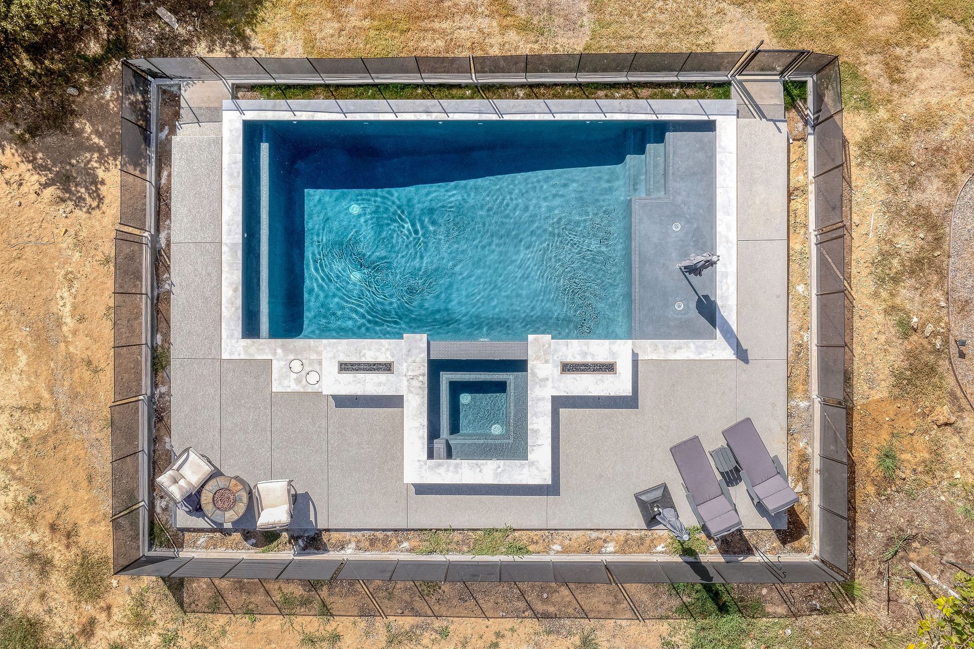 Aerial view of a backyard pool with a small hot tub, lounge chairs, and patio seating on a fenced deck.