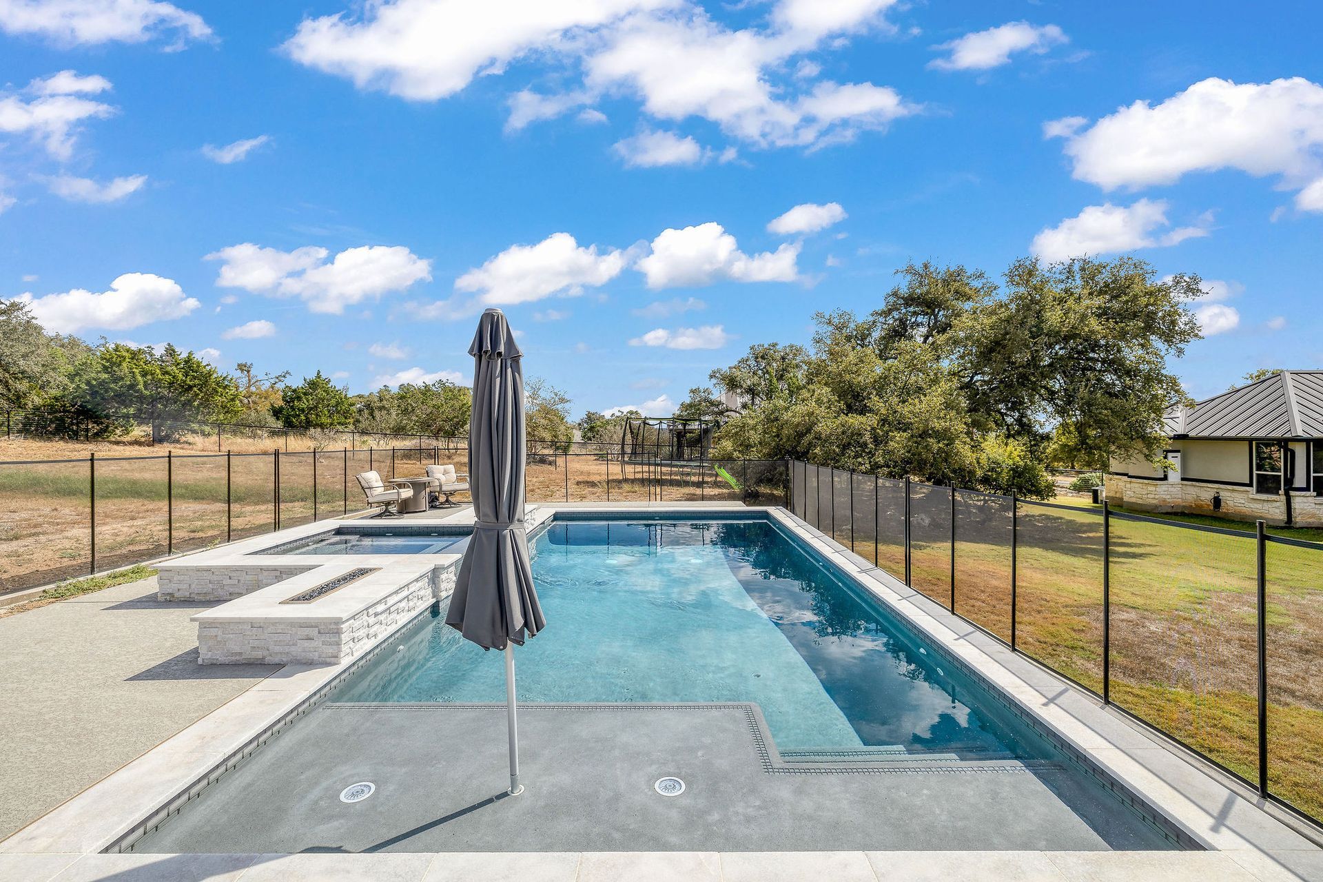 Backyard swimming pool with closed umbrella, patio, fence, and cloudy blue sky