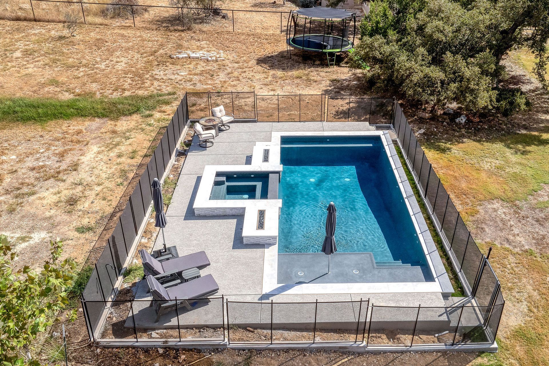 Small backyard pool with gray patio, lounge chairs, and fenced perimeter in a dry landscape
