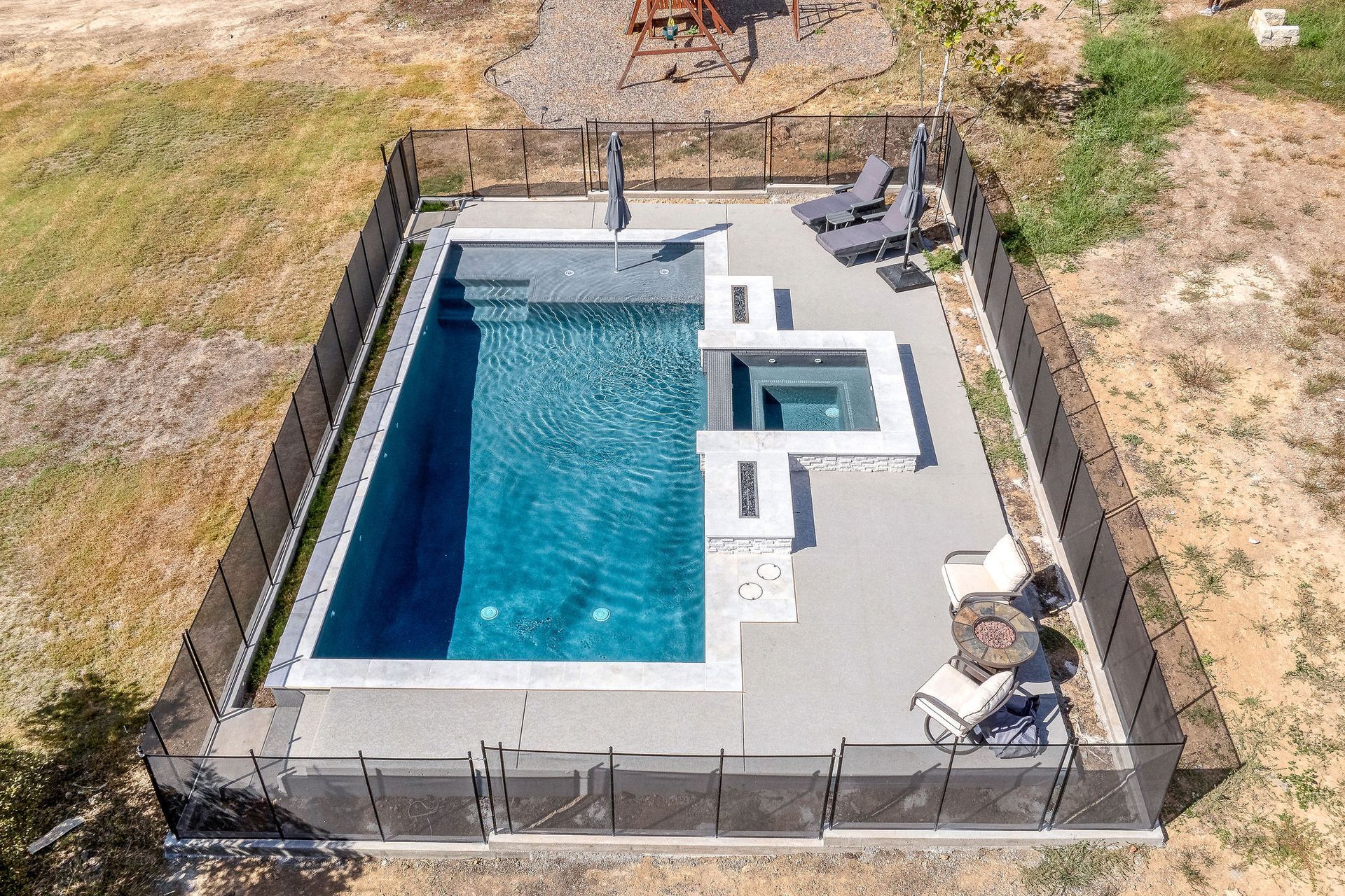Rectangular backyard pool with attached spa, surrounded by a fenced concrete patio in a dry landscape