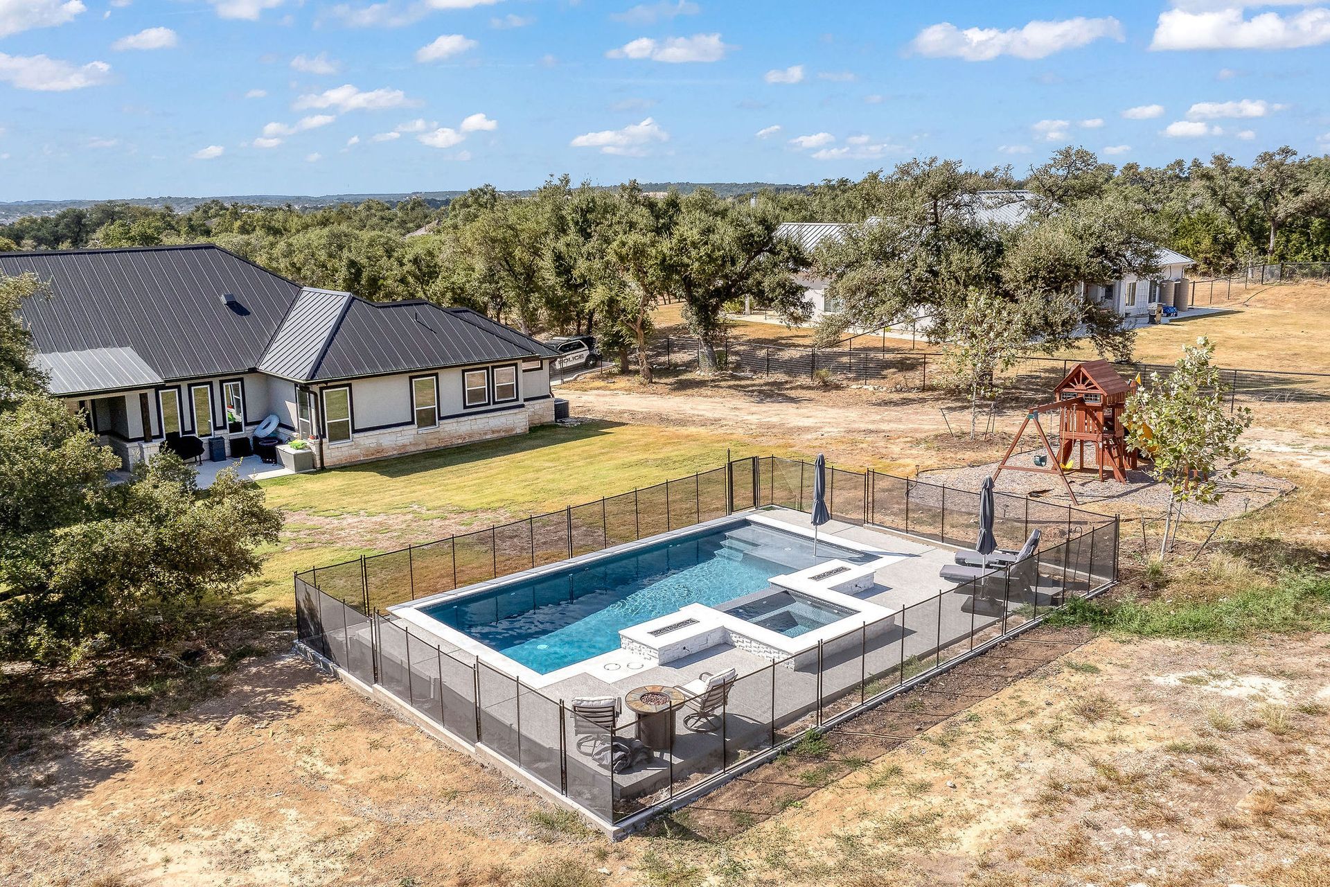 Backyard pool with patio furniture beside a house, surrounded by dry grass and trees under a blue sky
