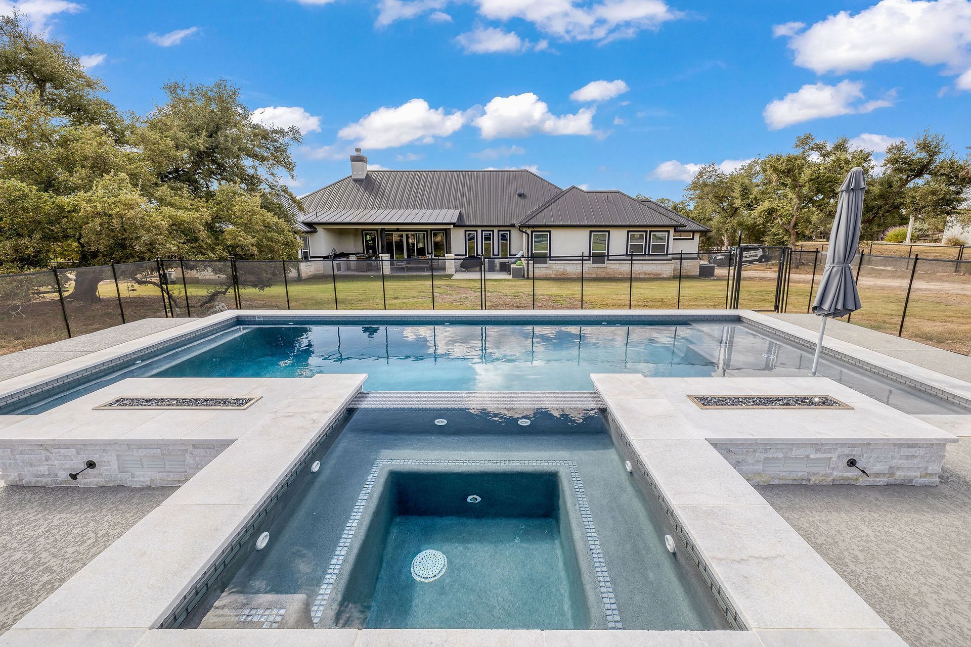 Modern backyard with a swimming pool, spa, and patio in front of a house under a blue sky