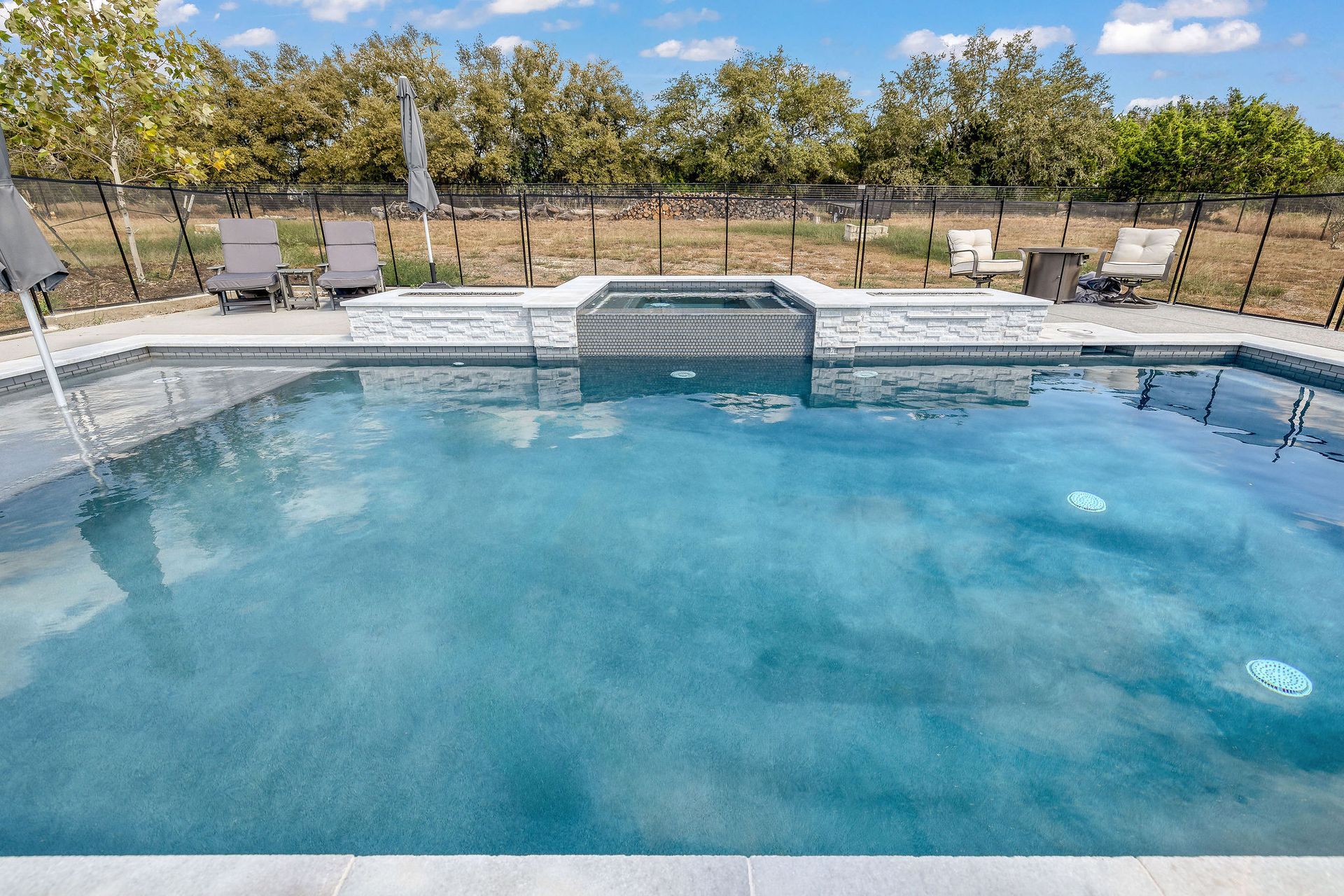 Backyard swimming pool with clear blue water, lounge chairs, and trees beyond a black fence