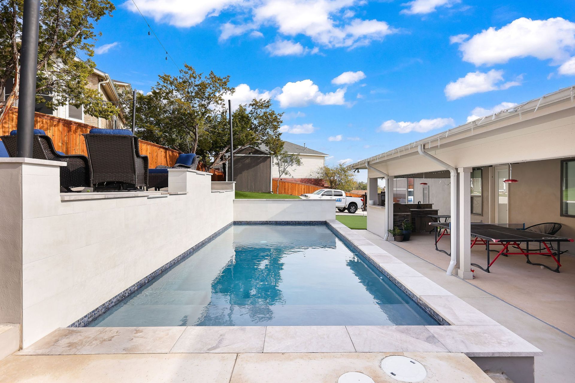 Modern backyard with a rectangular pool, white patio, and covered outdoor seating area under a blue sky.