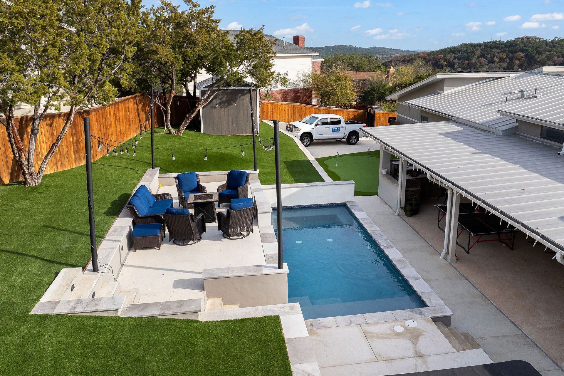 Backyard patio with blue chairs, dining table, lawn, and a long narrow swimming pool beside a covered deck.