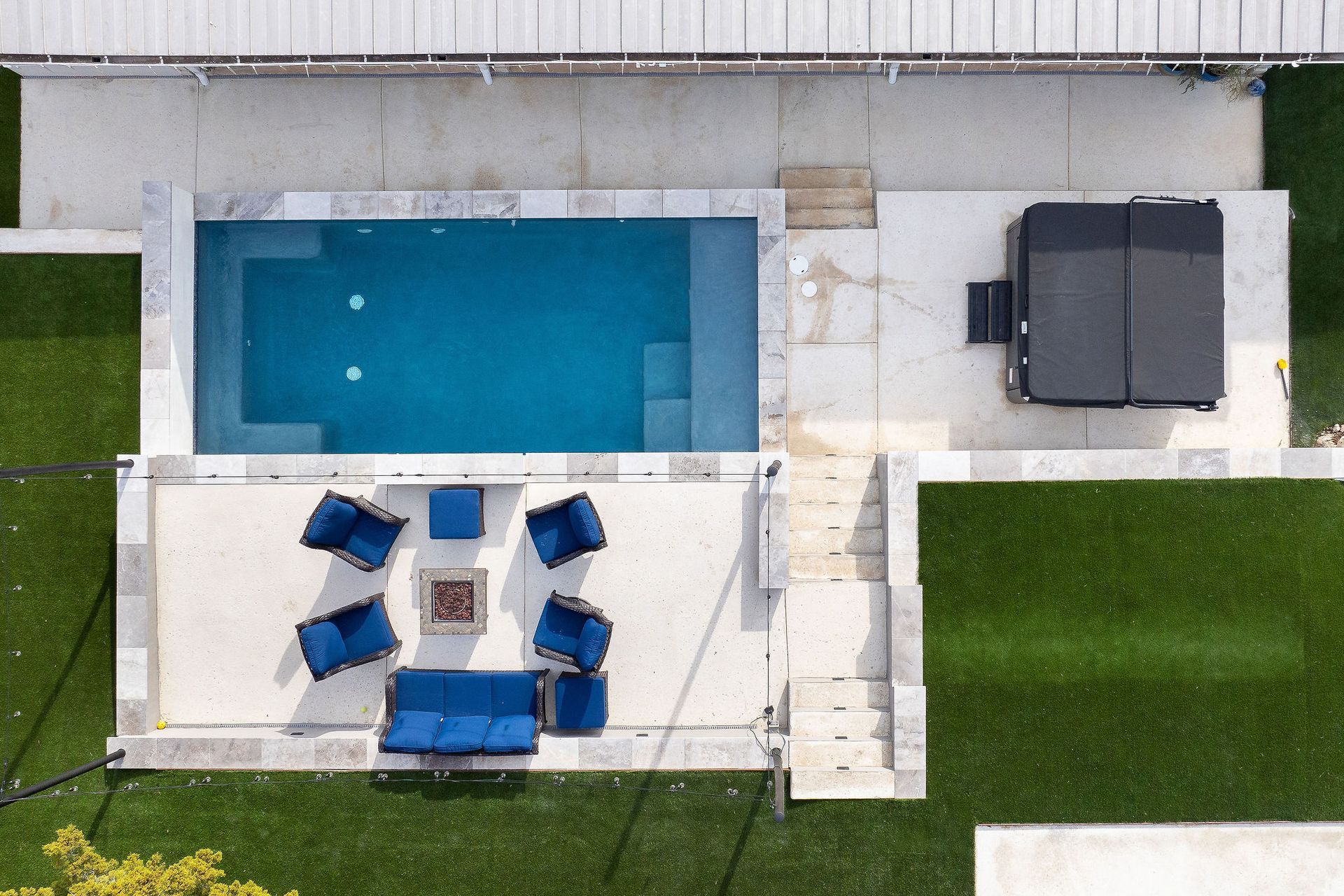 Aerial view of a backyard with a blue pool, patio seating, and a grill beside a house and lawn.