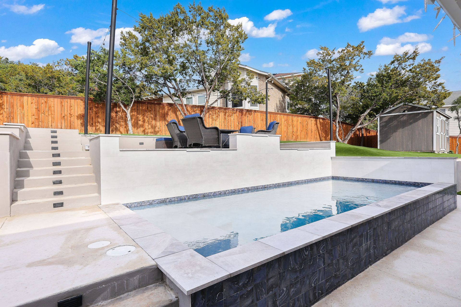 Modern backyard with a white patio, sunken pool, steps, and black outdoor seating under a blue sky