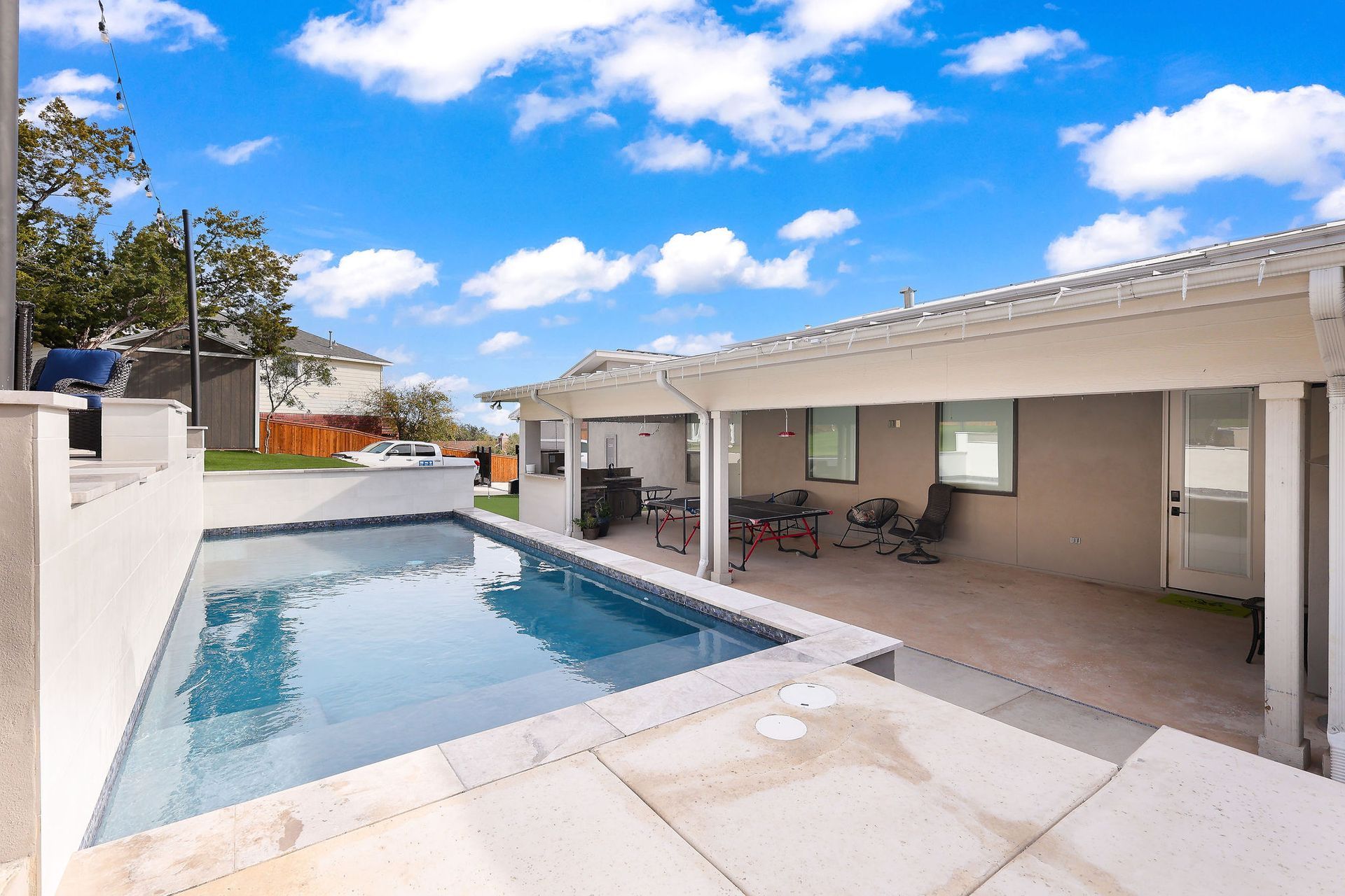 Backyard patio with a rectangular swimming pool, white walls, and a covered seating area under a blue sky