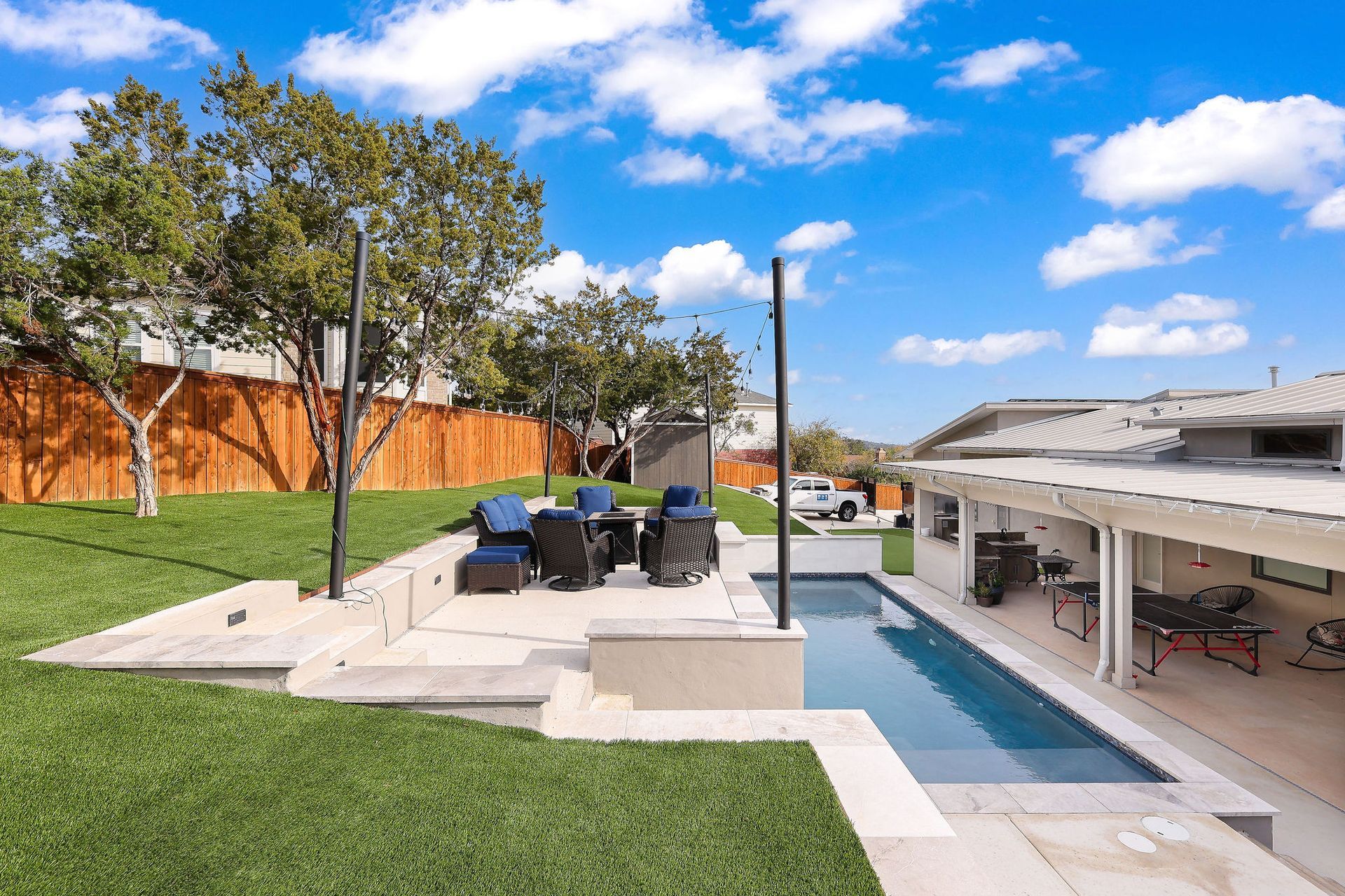 Modern backyard patio with dining table, lawn, pool, and covered outdoor seating under a bright blue sky