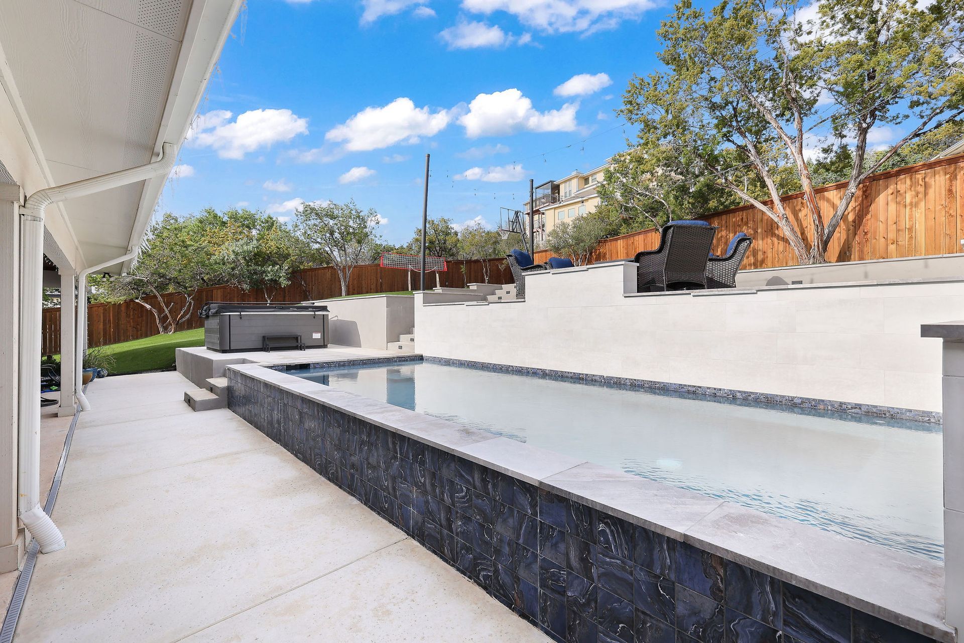 Modern backyard patio with a long rectangular pool, white deck, and lounge chairs under a blue sky
