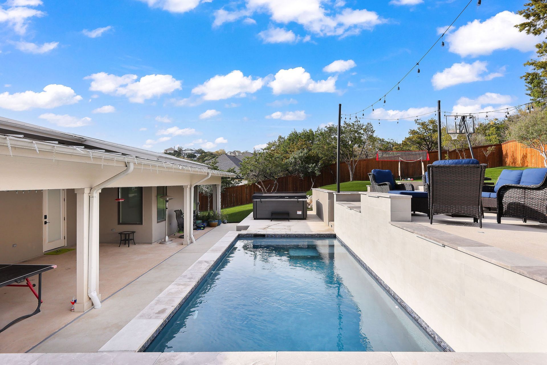 Backyard with a narrow swimming pool, white patio walls, and black lounge chairs under a blue sky.