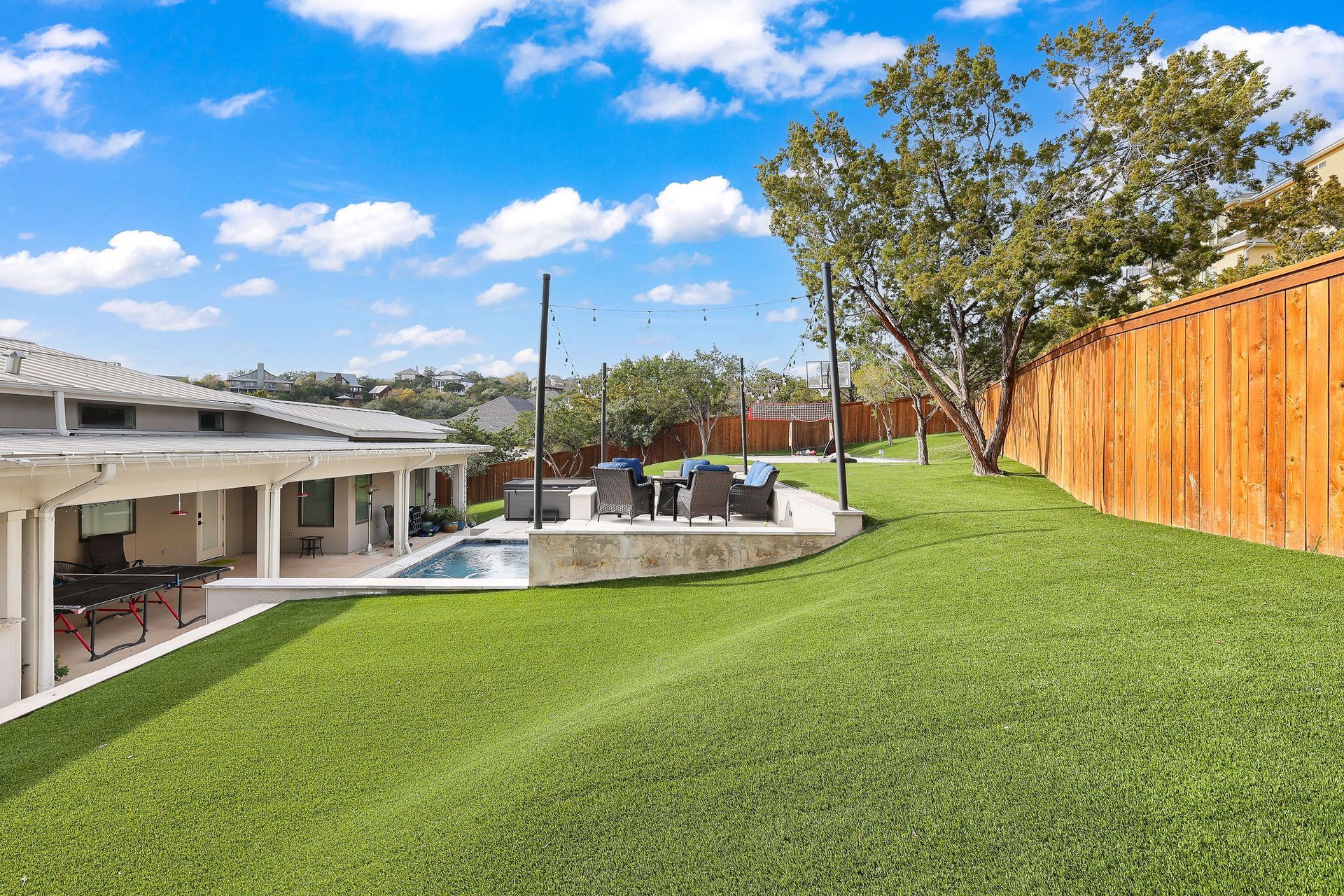 Backyard lawn beside a pool, patio, and wooden fence under a bright blue sky