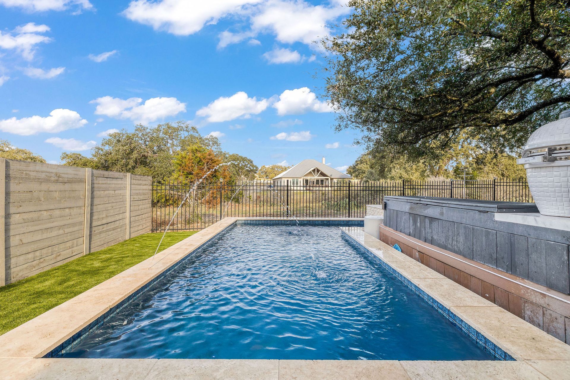 Backyard swimming pool with blue water, stone patio, fence, and a clear sky
