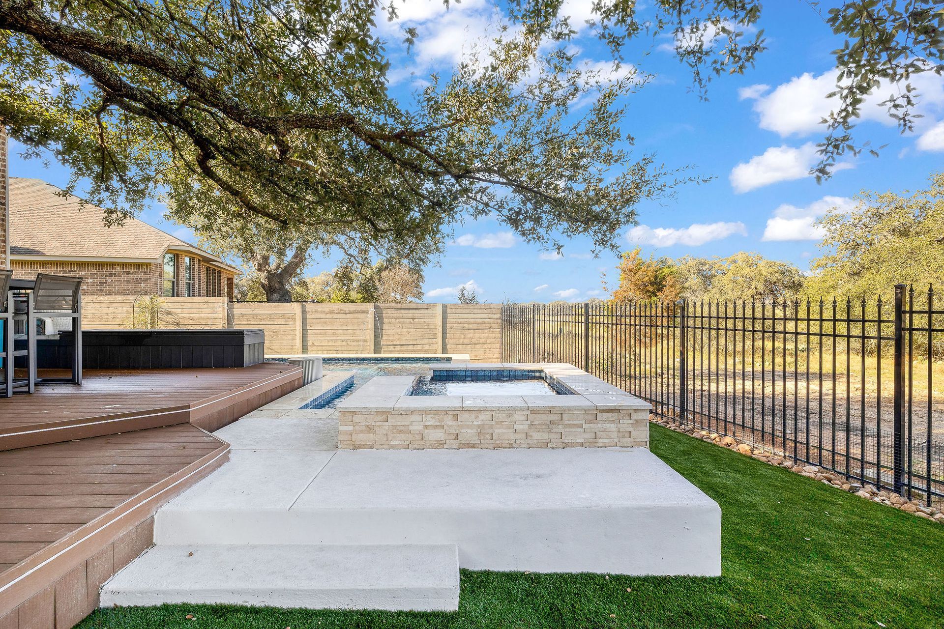 Backyard patio with a raised hot tub, wooden deck, green lawn, and black metal fence under a blue sky