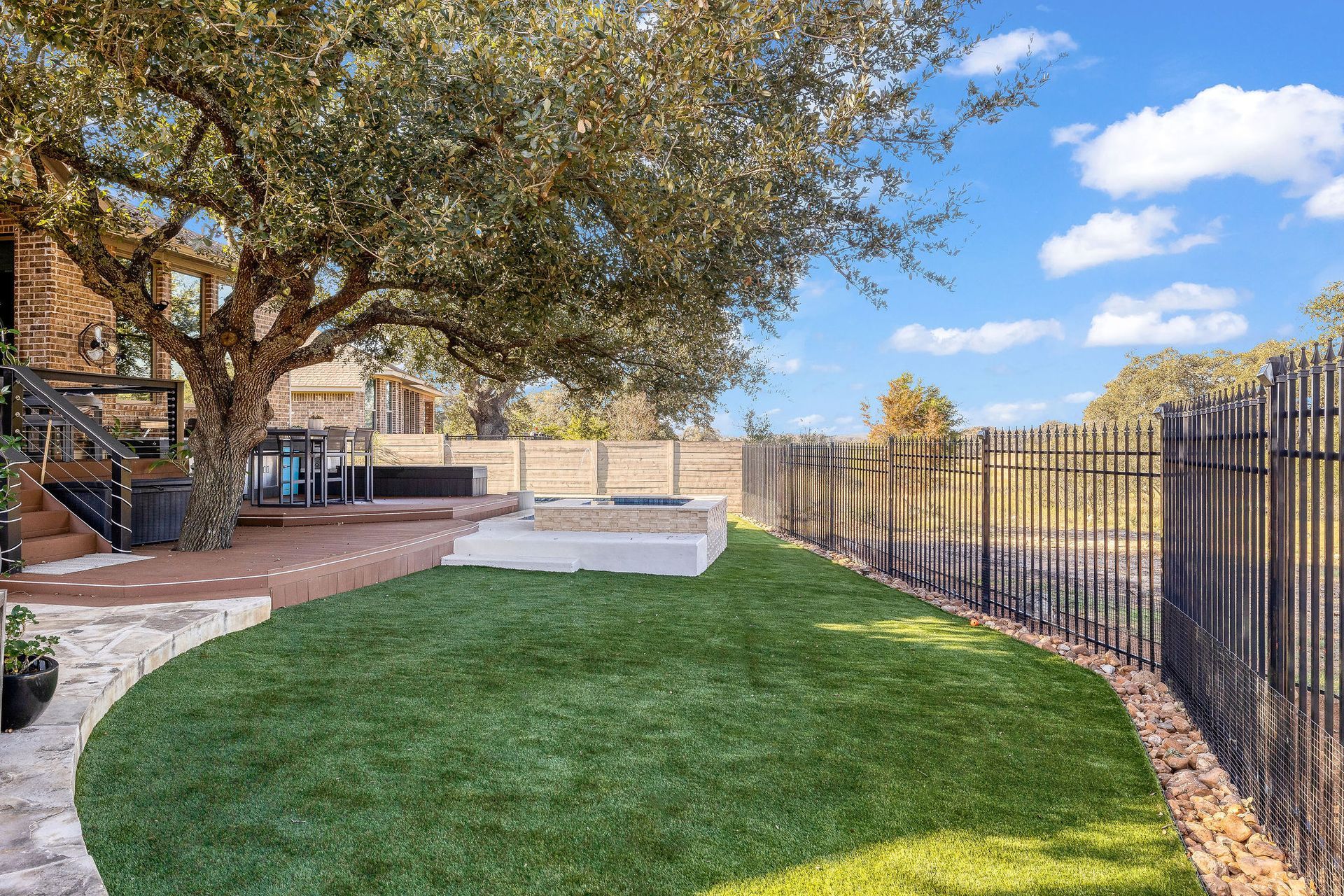 Backyard with green lawn, stone patio, large tree, and black metal fence under a blue sky