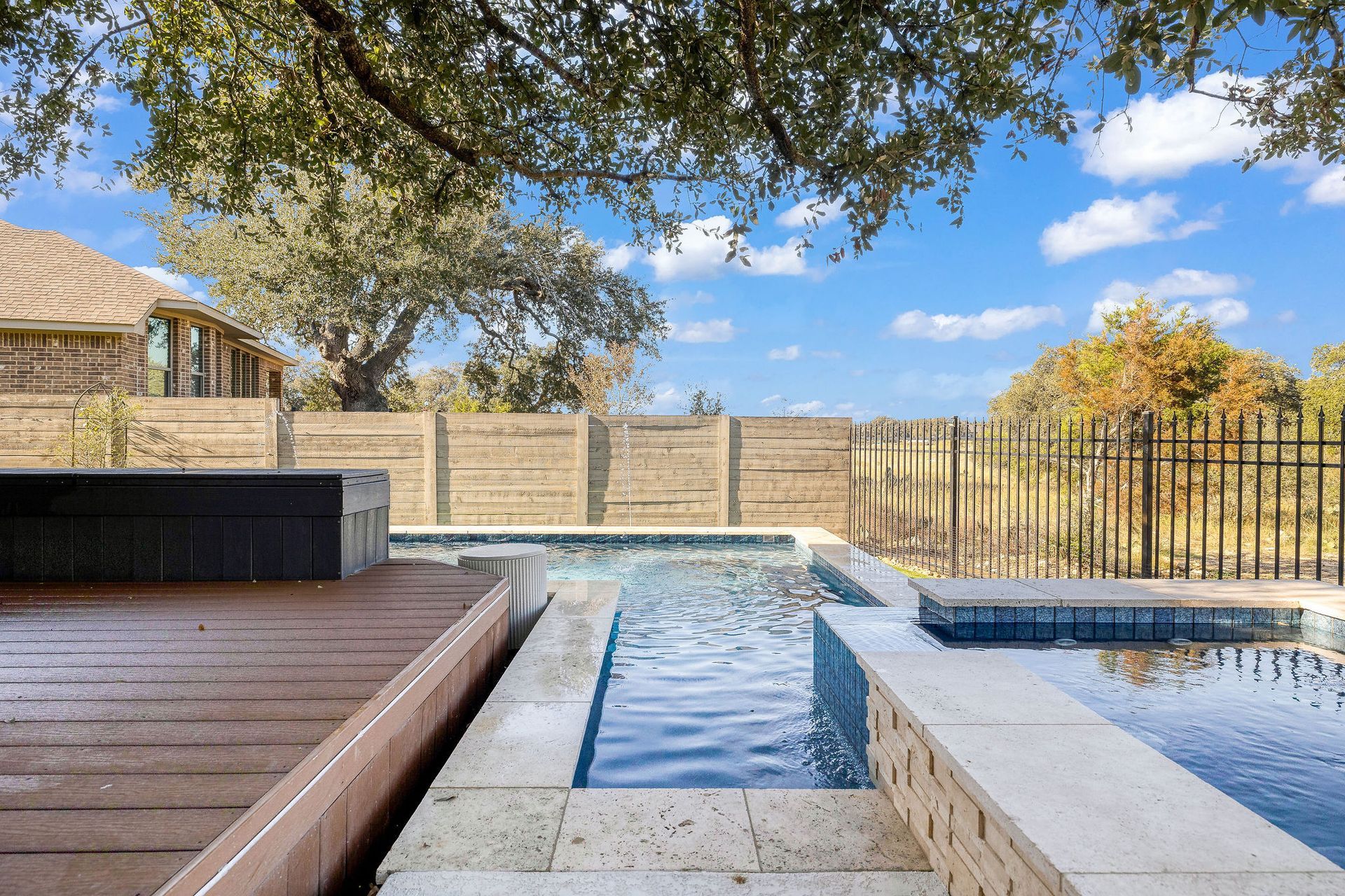 Backyard pool and hot tub beside a wooden deck and fence under a blue sky