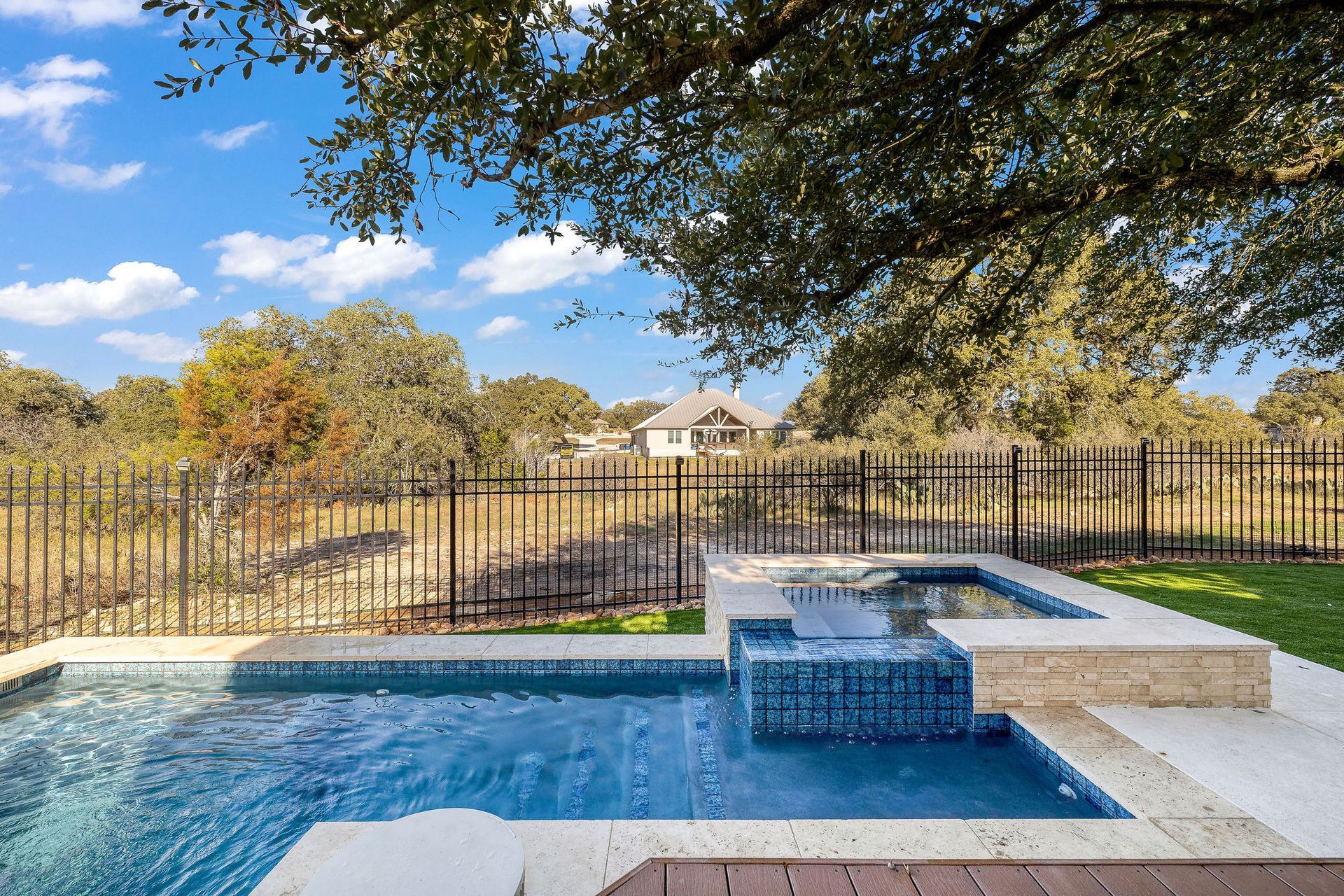 Backyard pool with blue water, stone deck, and fenced lawn under a tree