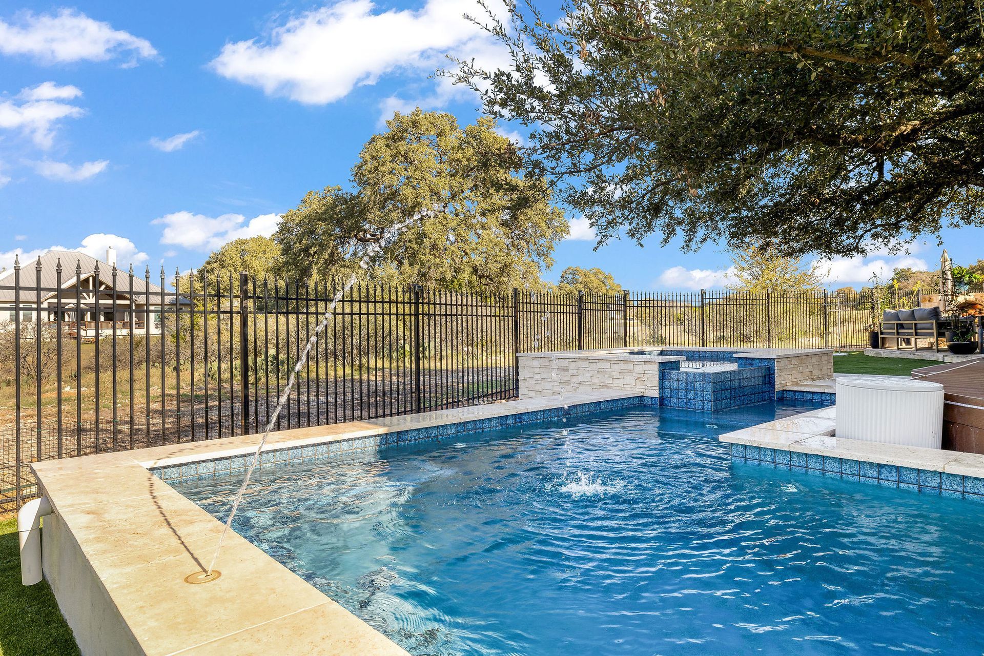 Backyard swimming pool with blue water, stone deck, black fence, and trees under a sunny sky
