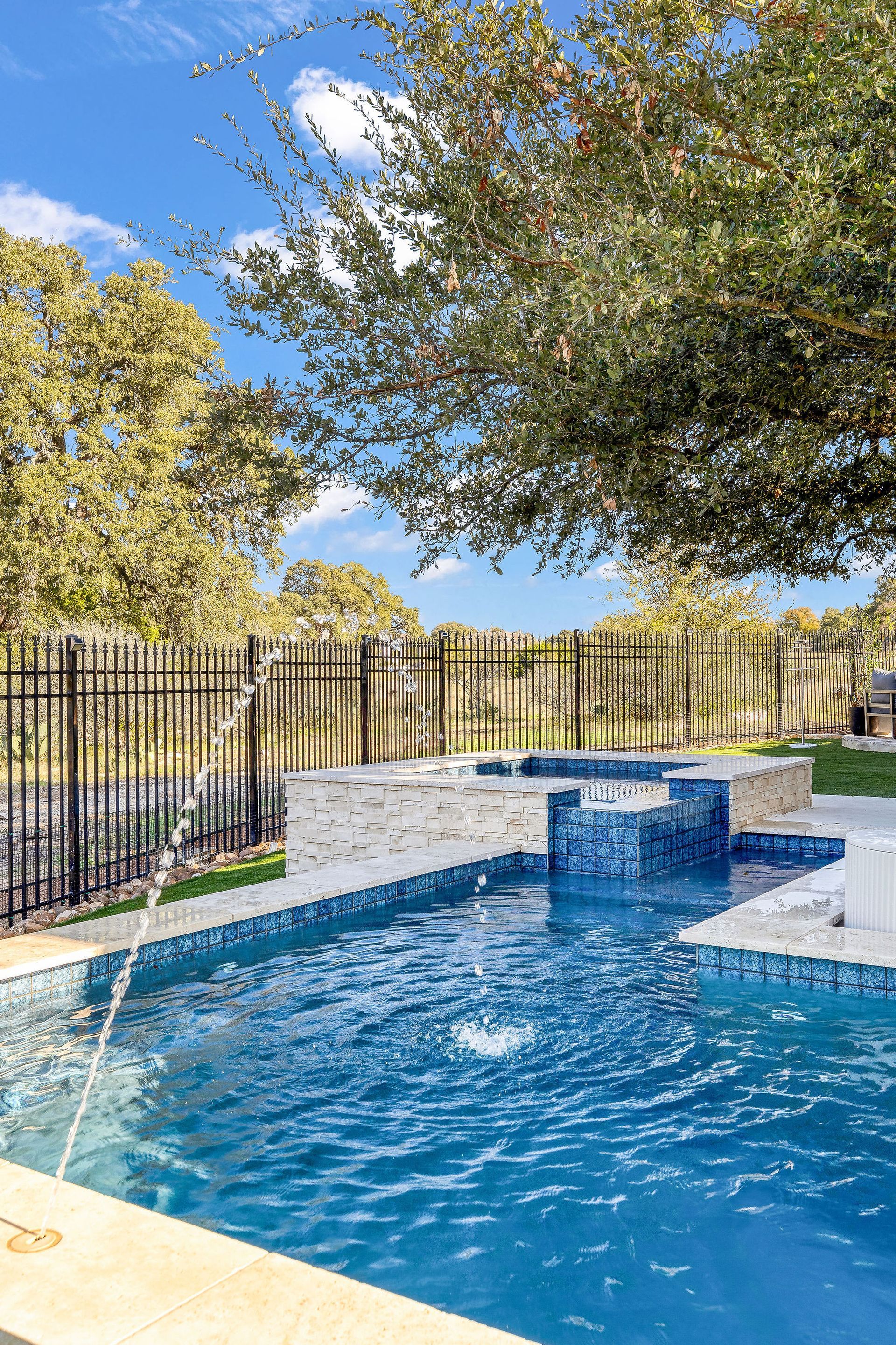 Backyard pool with blue water, stone deck, black fence, and trees under a clear sky