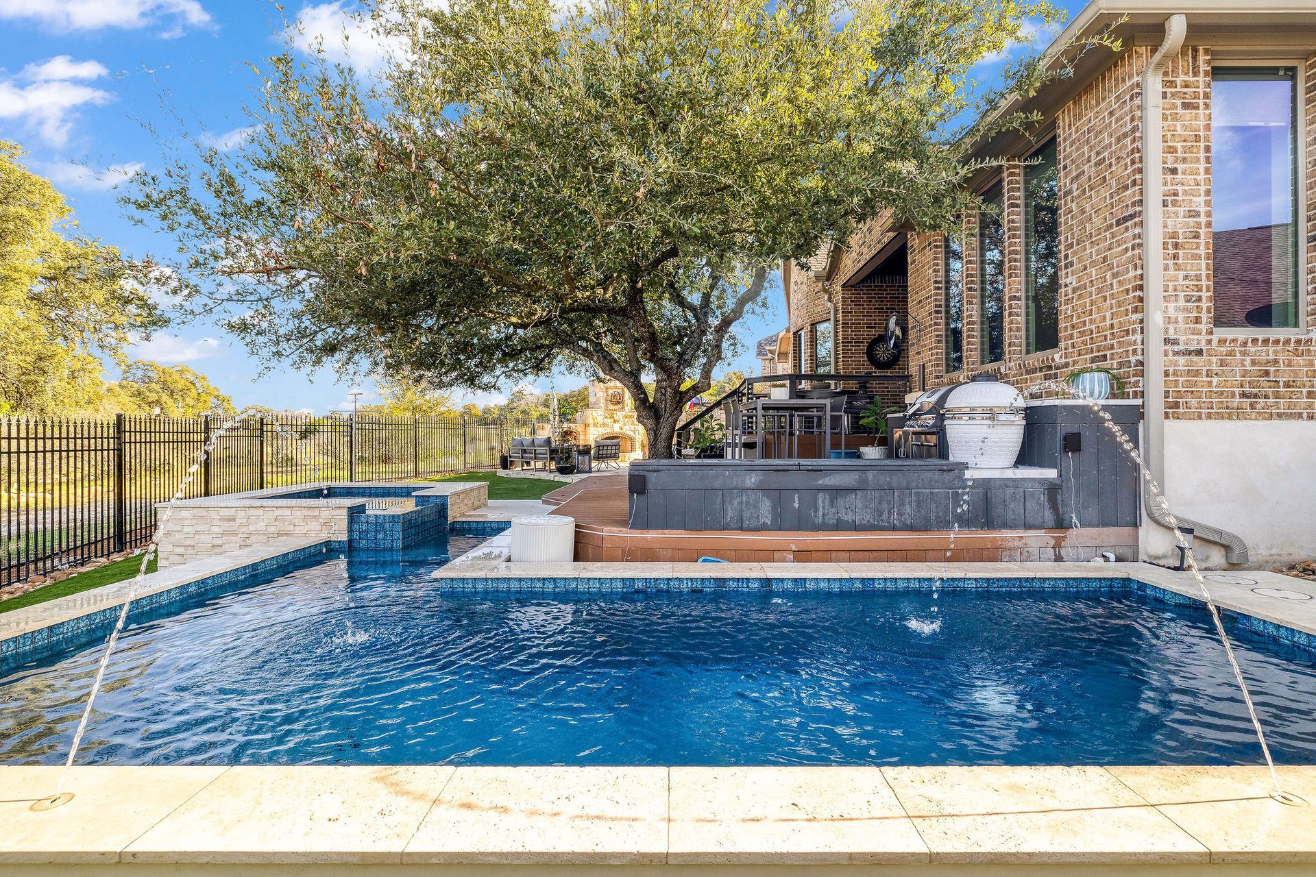 Backyard pool with blue water, stone patio, brick house, and a large tree overhead