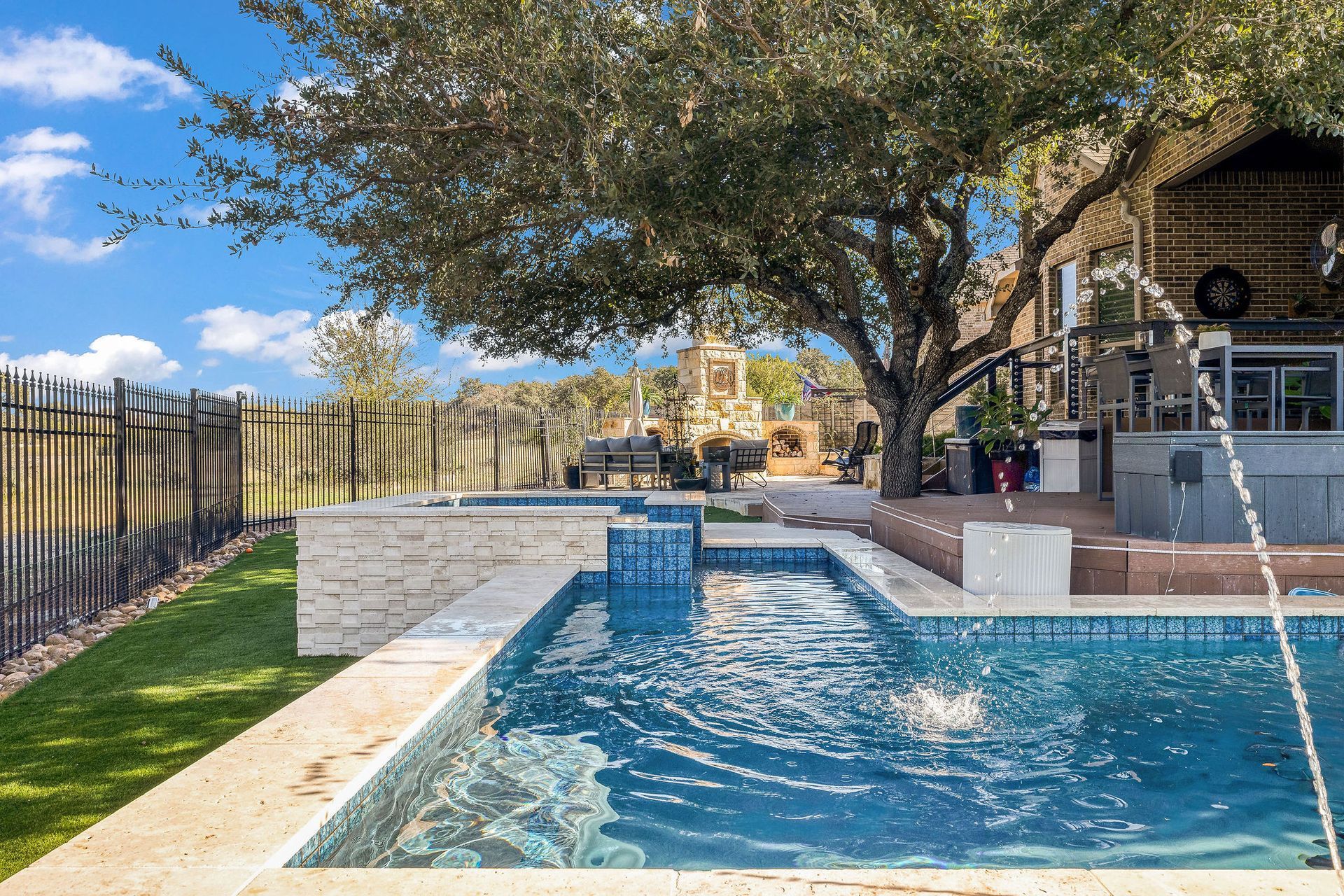 Backyard pool with water feature, stone deck, shaded trees, and patio seating under a blue sky