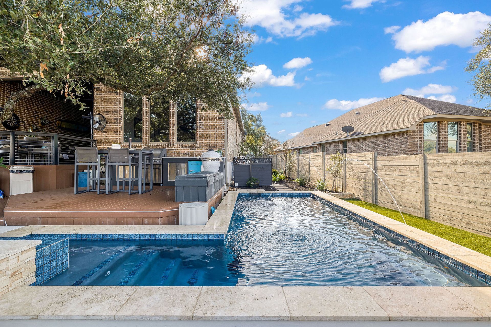 Backyard with a rectangular swimming pool, patio seating, and a brick house under a blue sky.