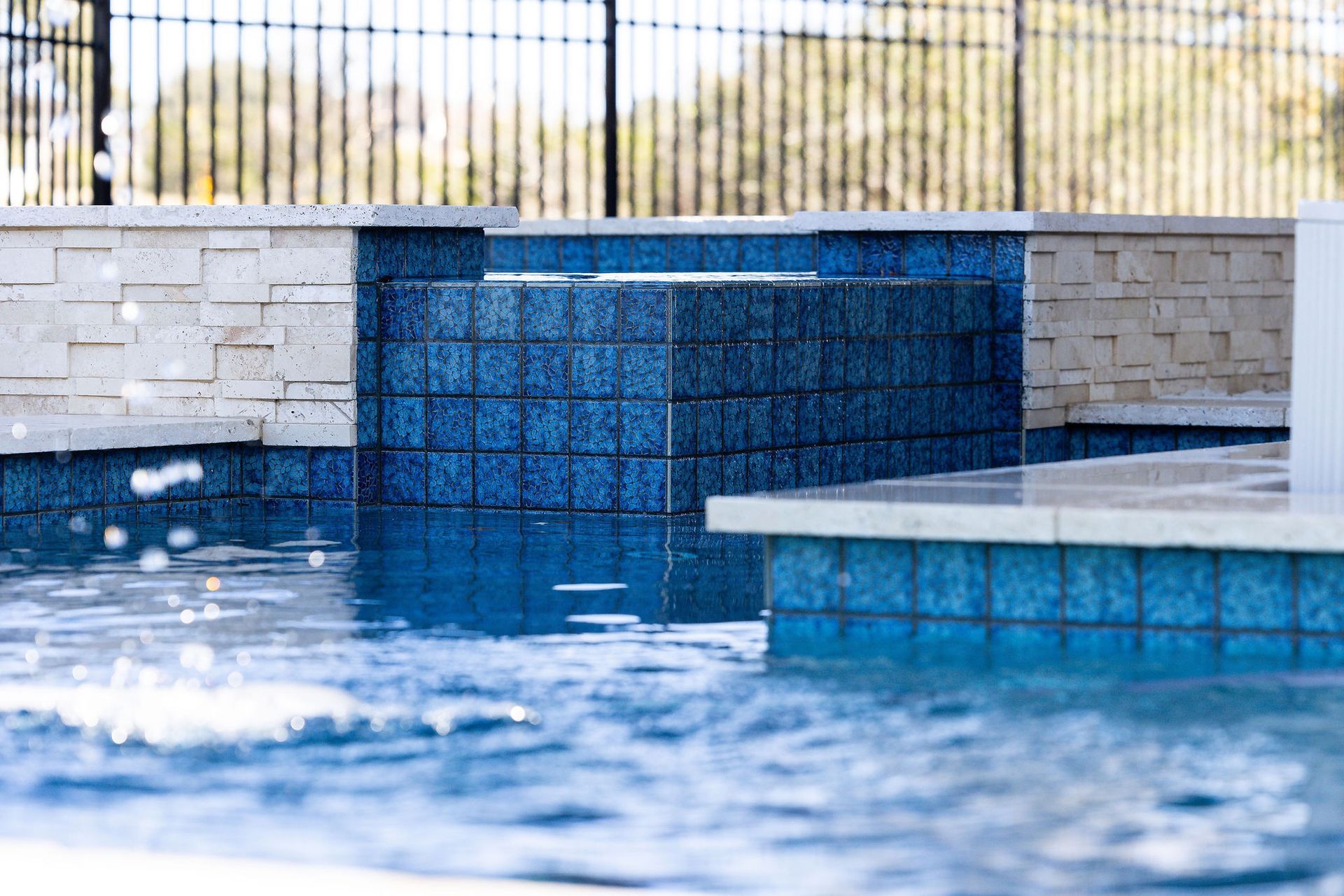 Blue-tiled swimming pool with a water feature and stone coping by a fence