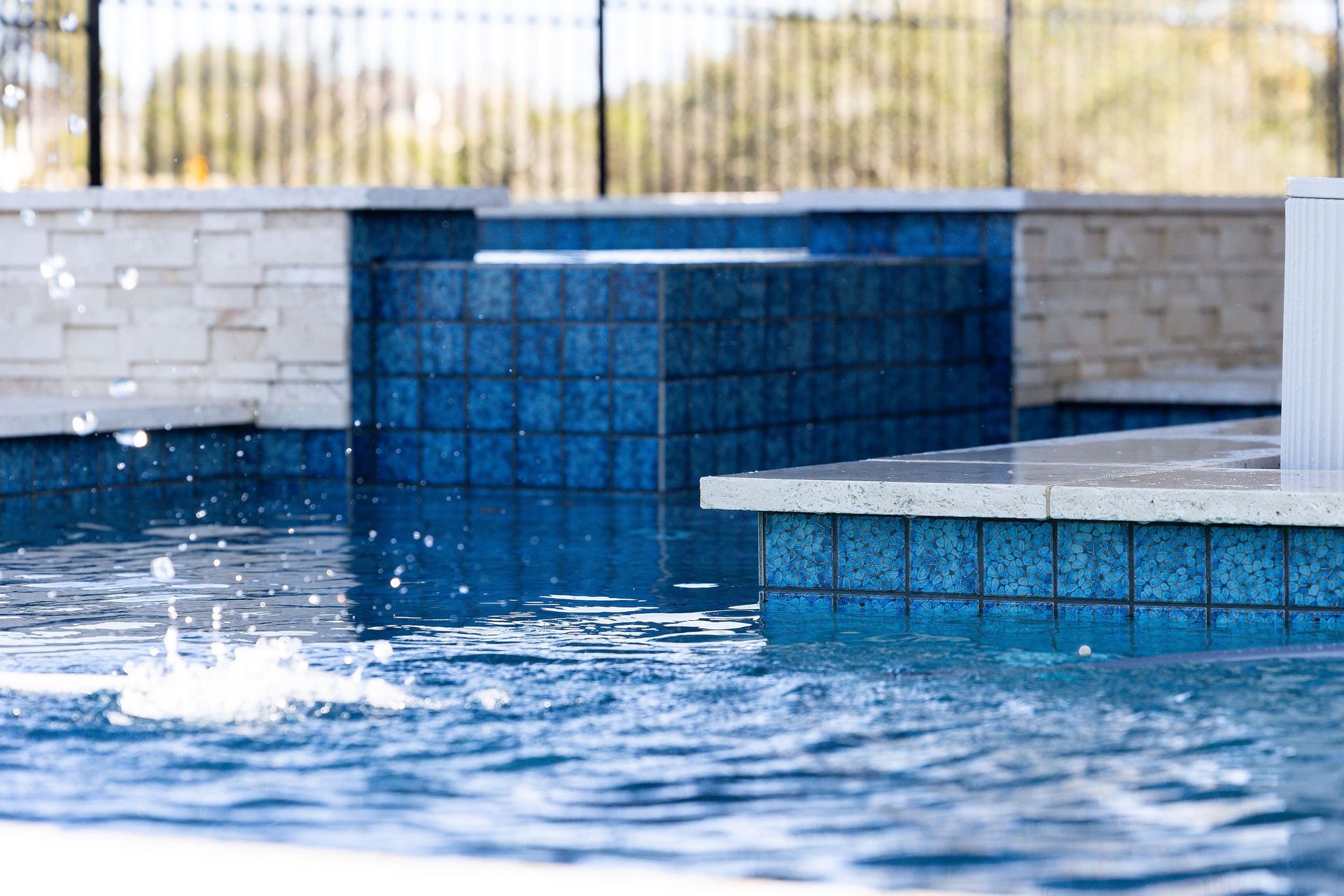 Blue tiled pool with water splashing near a stone ledge and raised wall