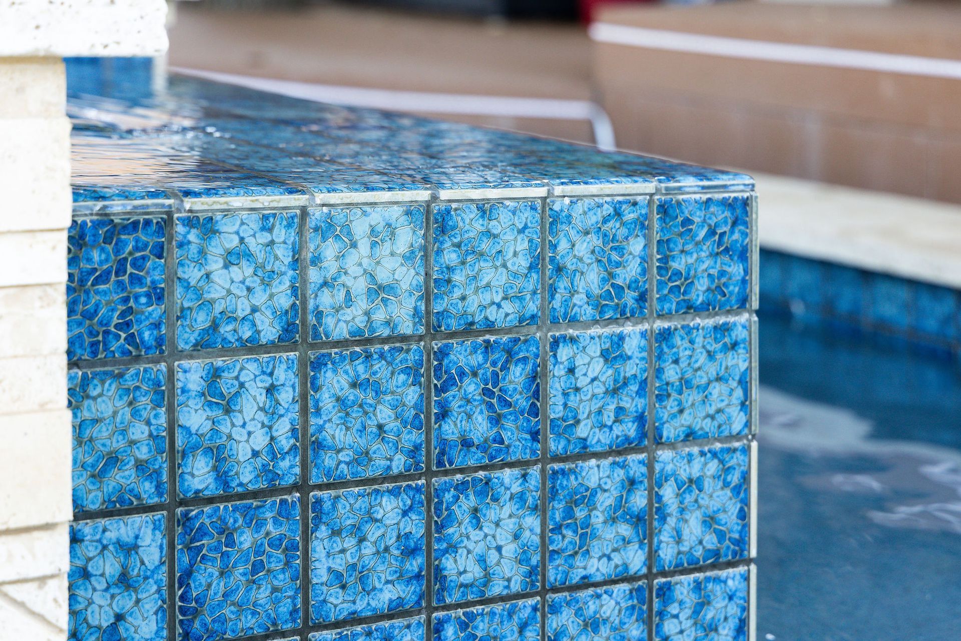 Blue tiled pool edge beside water with a blurred white column and deck in the background
