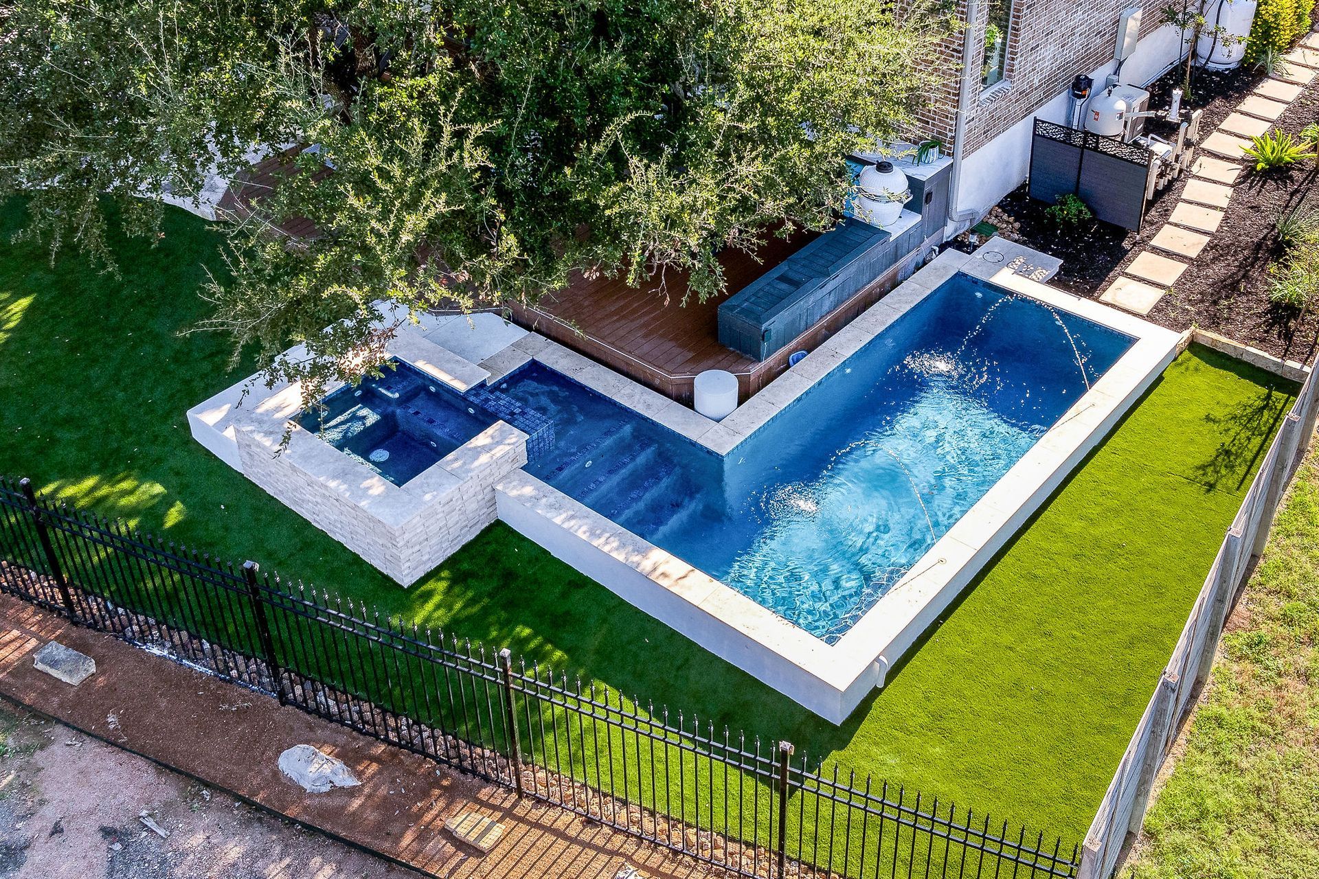 Aerial view of a small blue swimming pool beside a patio, green lawn, and black fence.