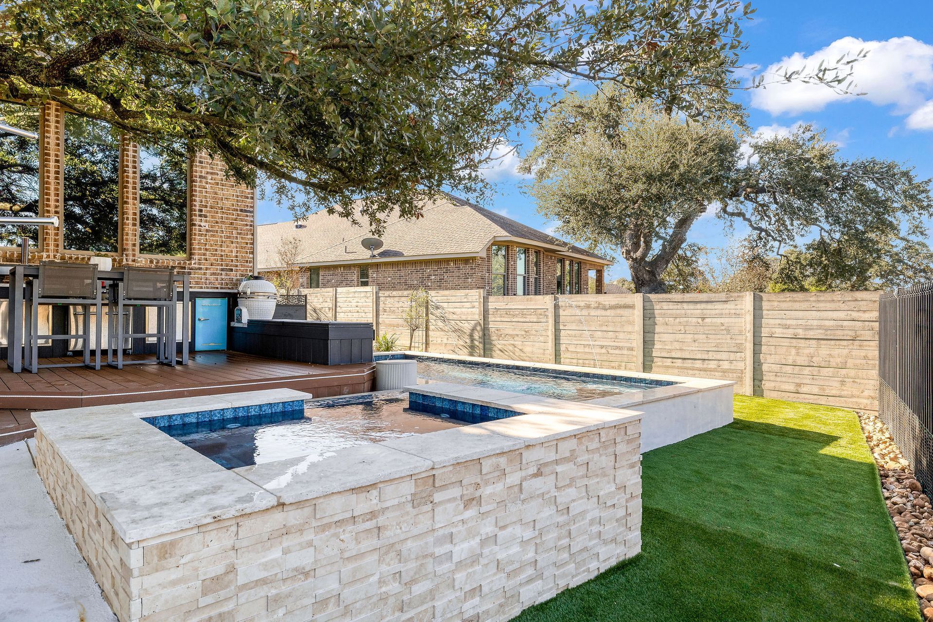 Backyard patio with a sunken fire pit, stone seating, and a fenced lawn under trees