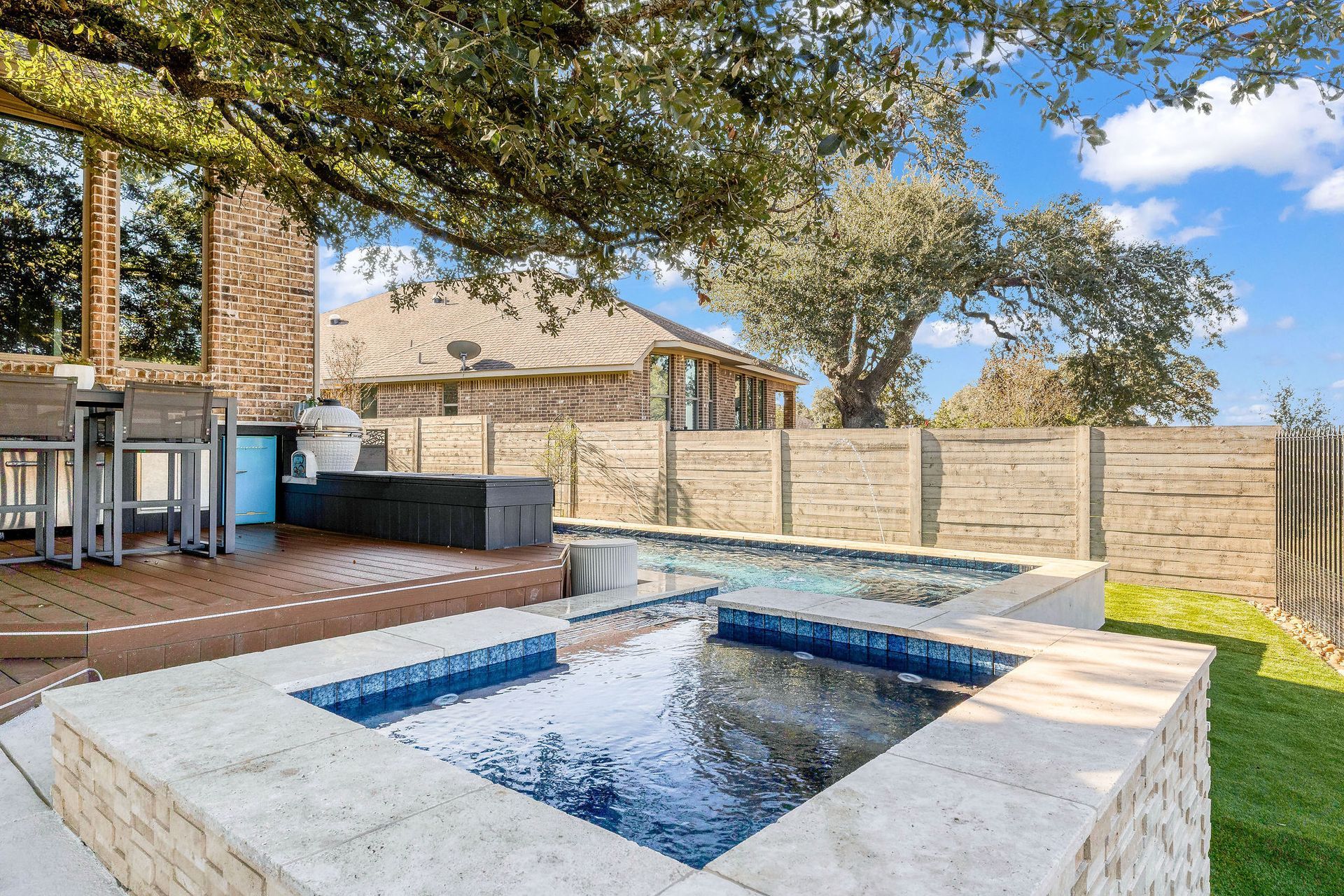 Backyard patio with a small square hot tub, deck seating, stone wall, and trees overhead