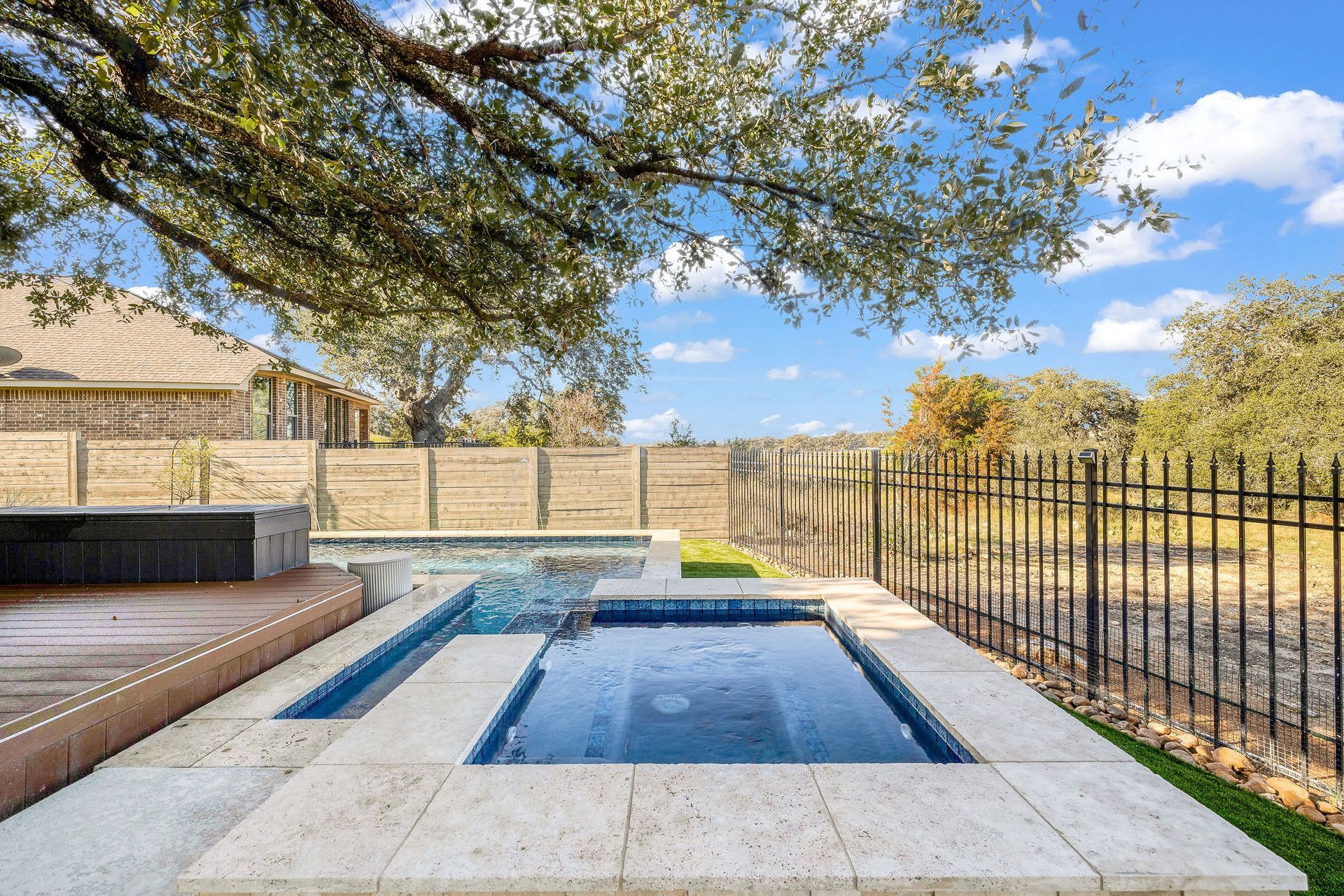 Backyard pool with a built-in spa, stone patio, and black metal fence under a leafy tree