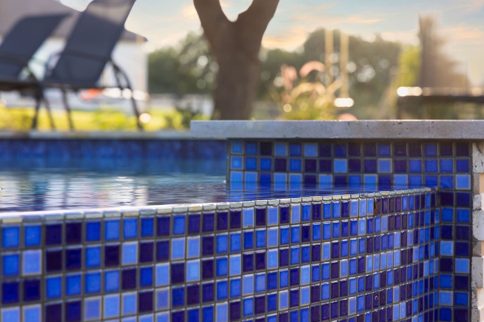 Blue mosaic-tiled pool edge with water and a blurred lounge chair in the background