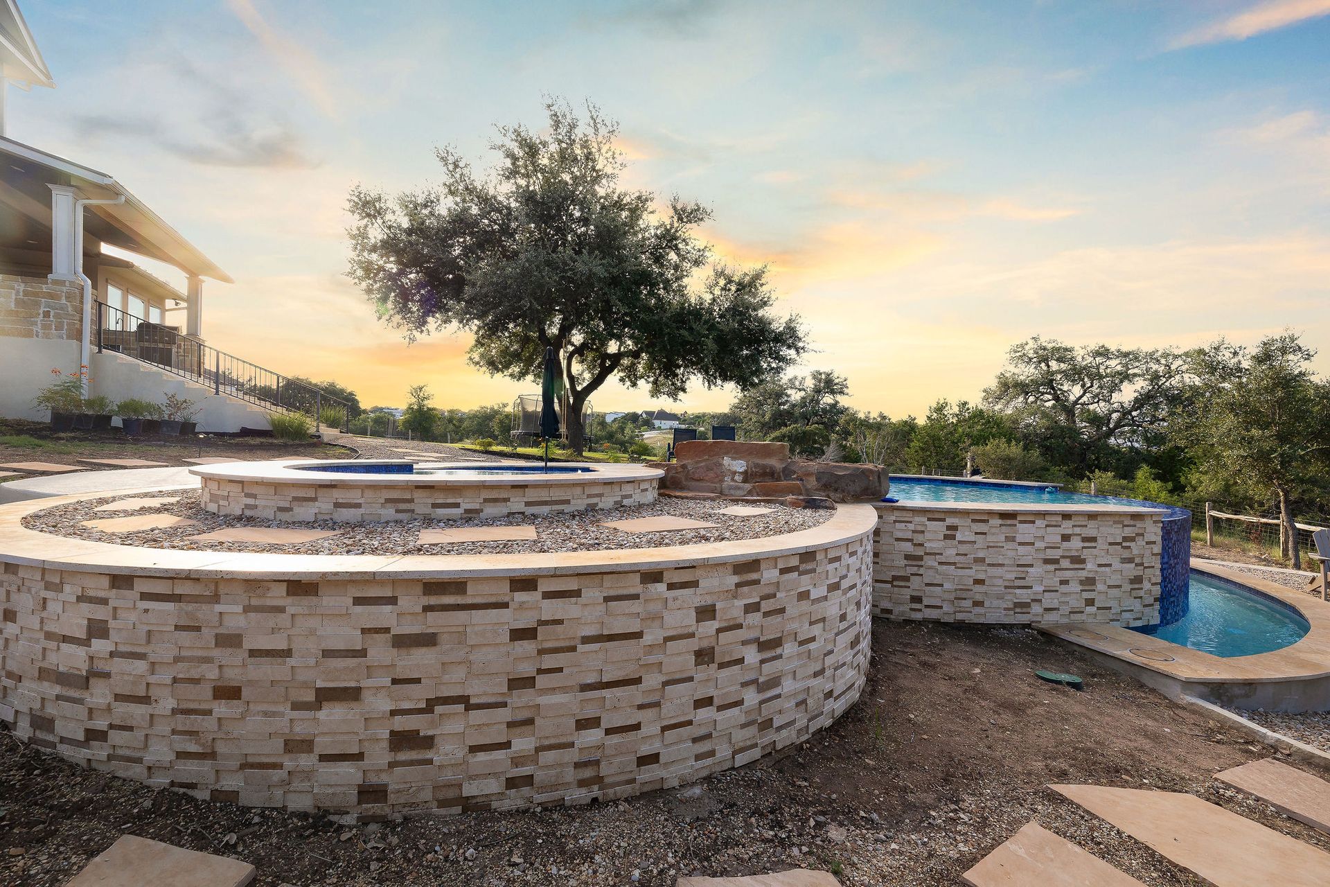 Backyard patio with circular stone fire pit, pool, and a tree at sunset