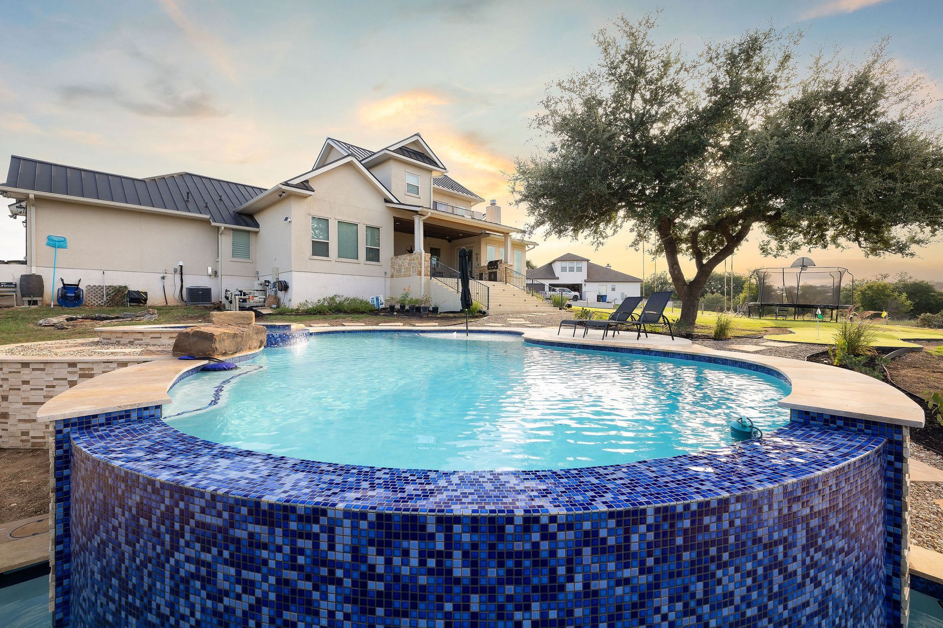 Backyard pool with blue mosaic tile edge beside a house and tree at sunset