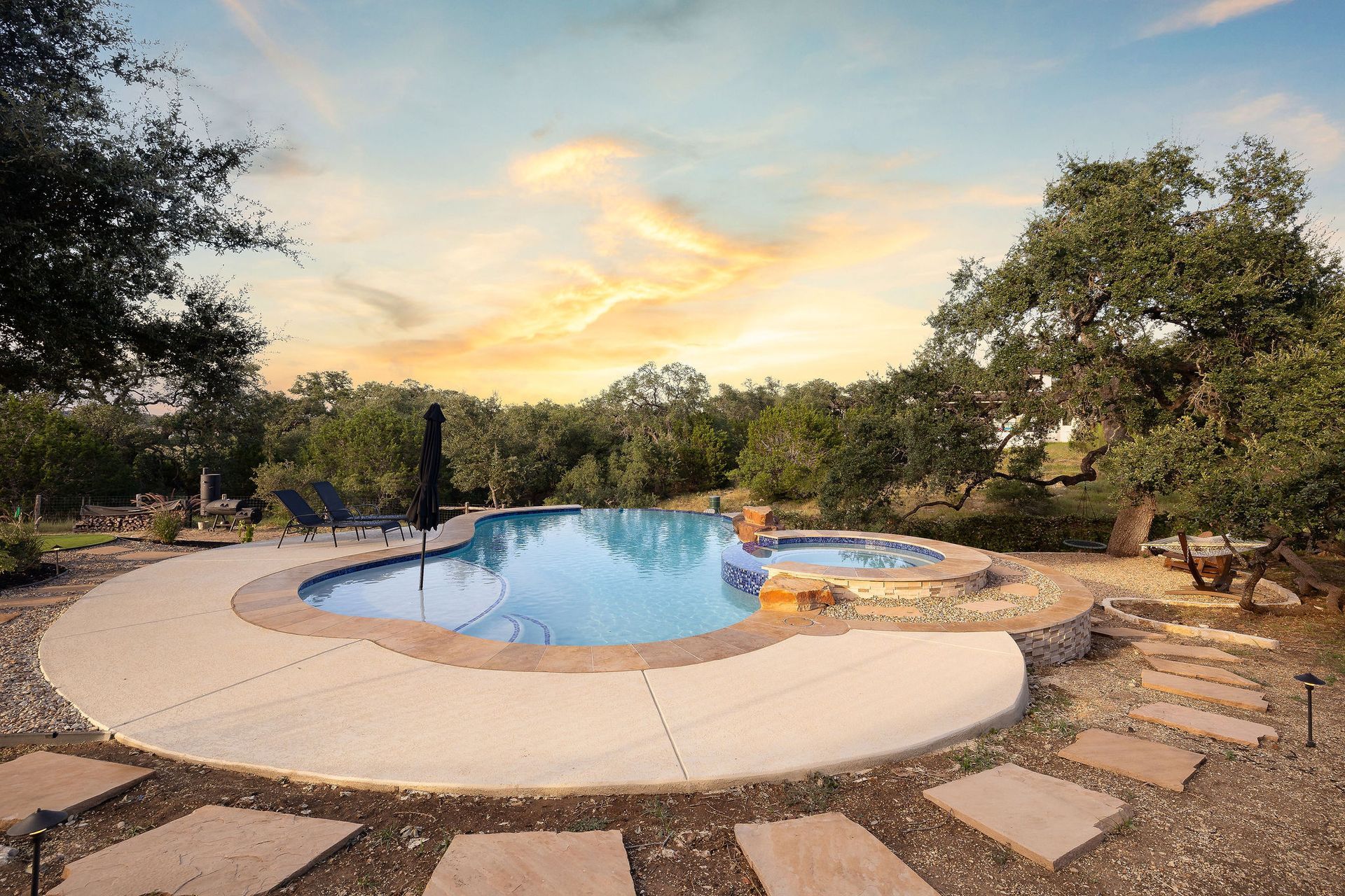 Backyard pool at sunset with stone patio and trees surrounding it