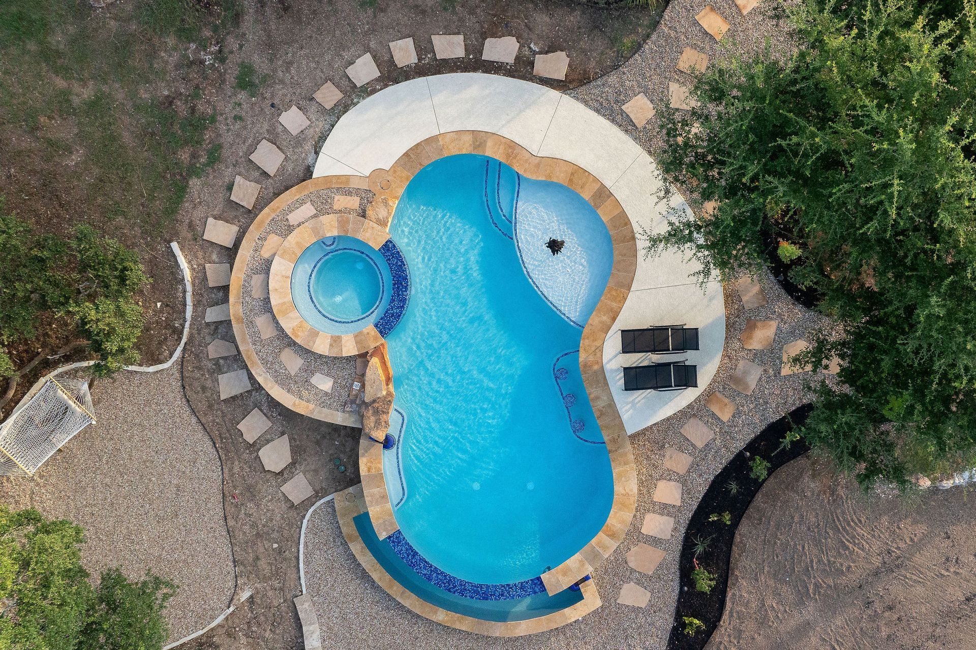 Top-down view of a curvy backyard pool with stone paving, a small spa, and surrounding greenery