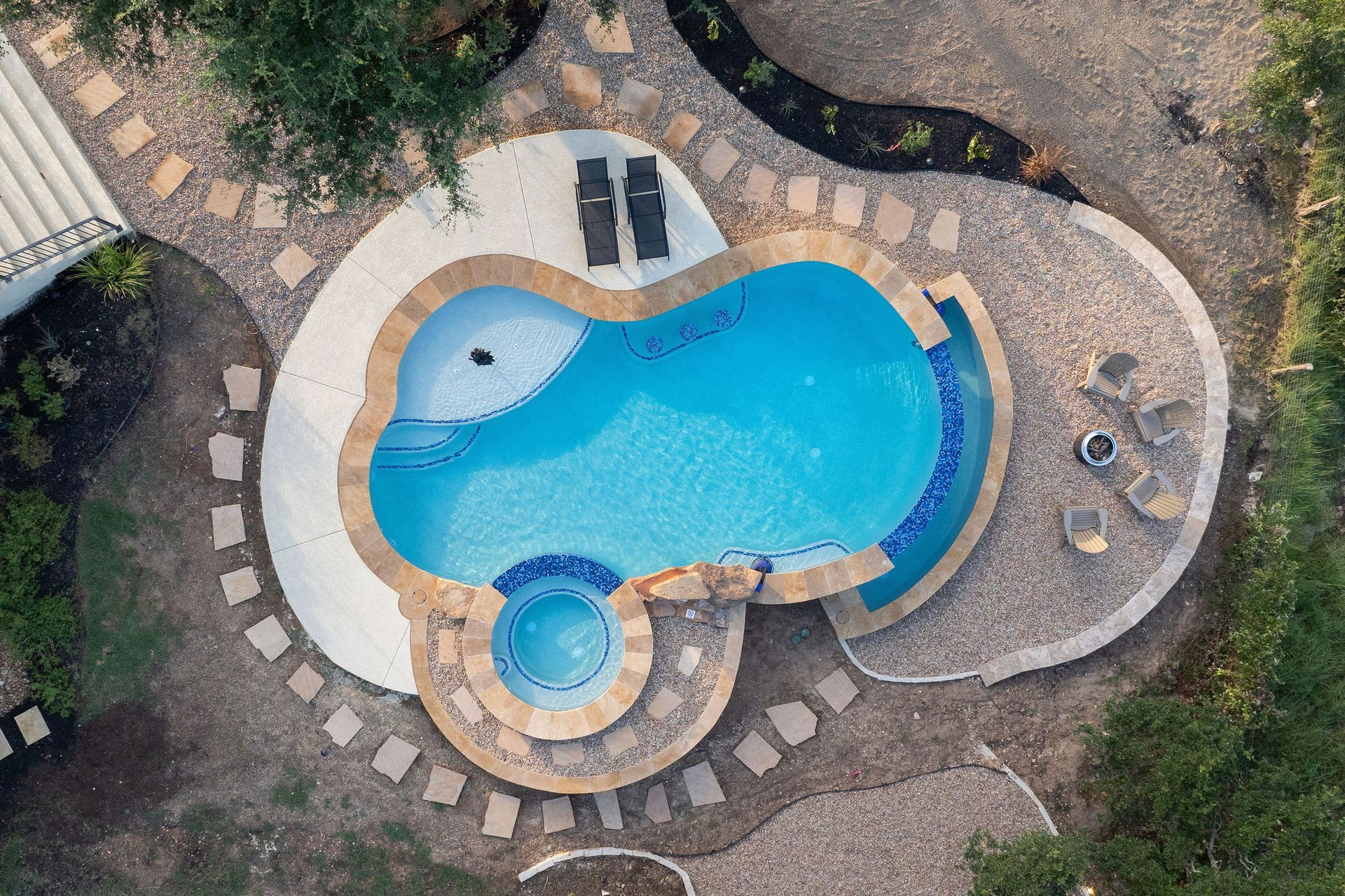 Top-down view of a curvy backyard pool with stone paving, a small spa, and surrounding greenery