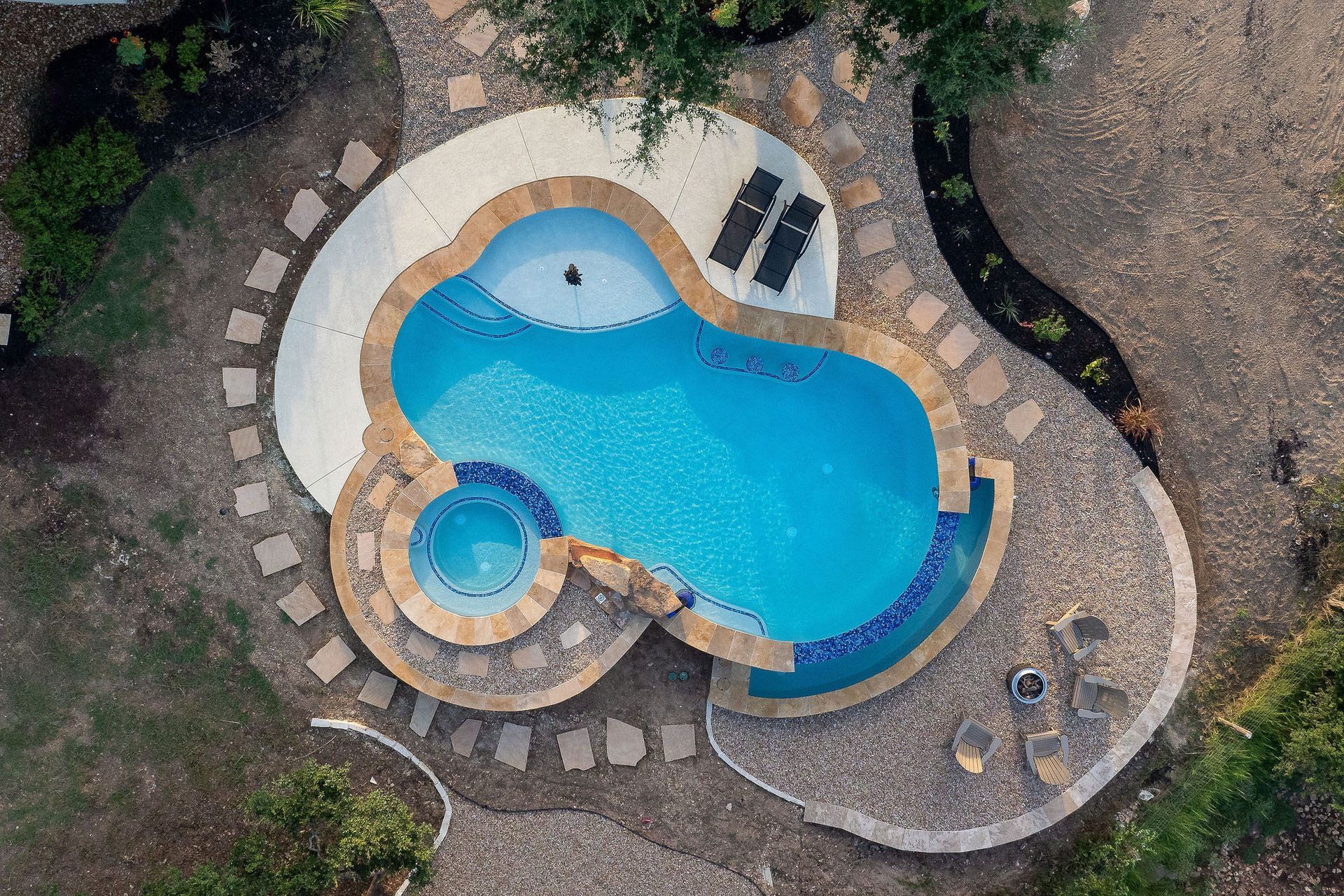 Top-down view of a curvy backyard pool with stone paving, a small spa, and surrounding greenery