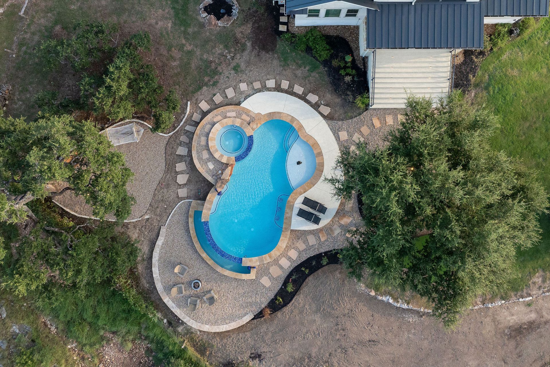 Top-down view of a curvy backyard pool with stone paving, a small spa, and surrounding greenery