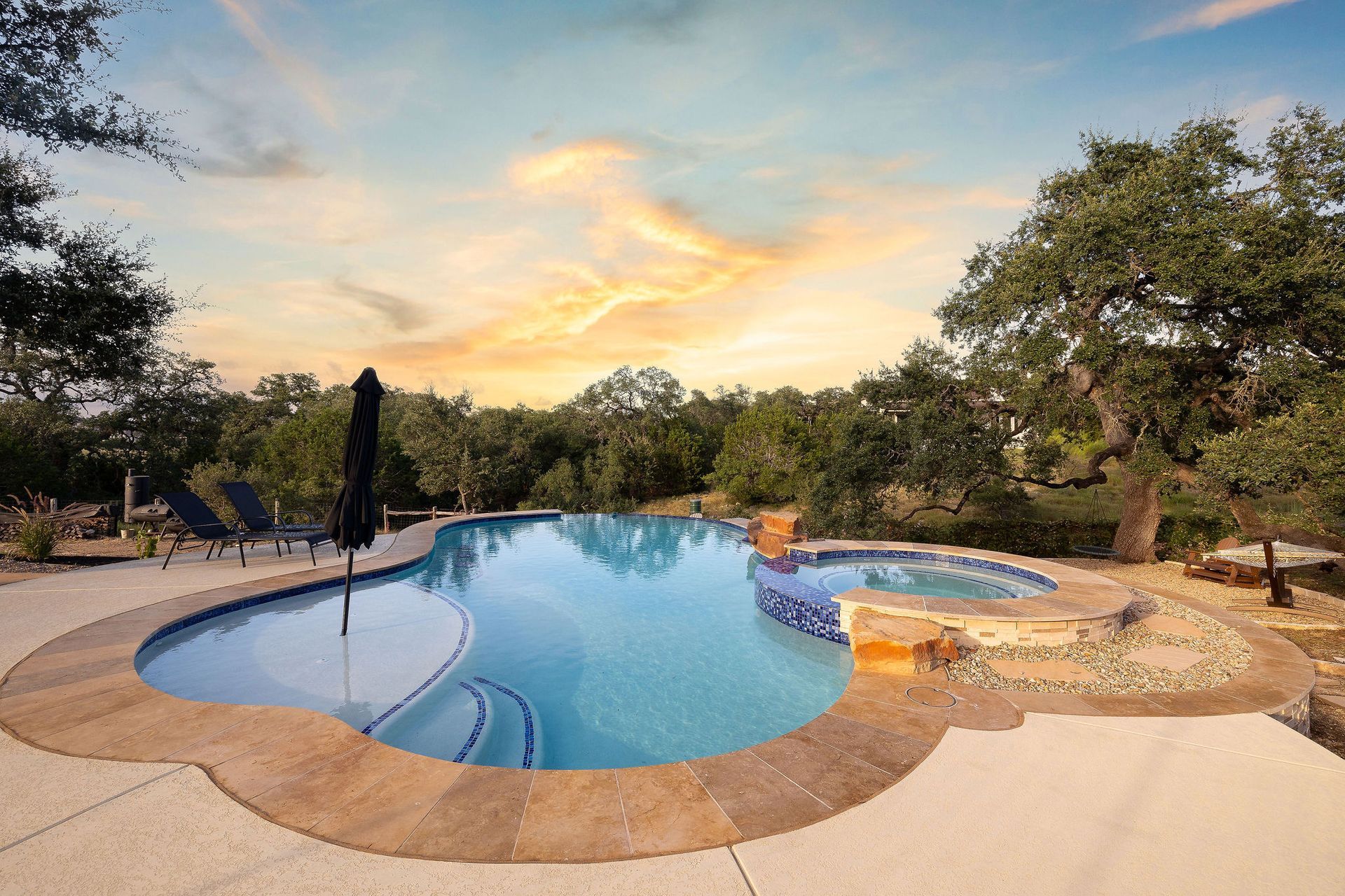 Backyard swimming pool at sunset with lounge chairs, trees, and a hot tub