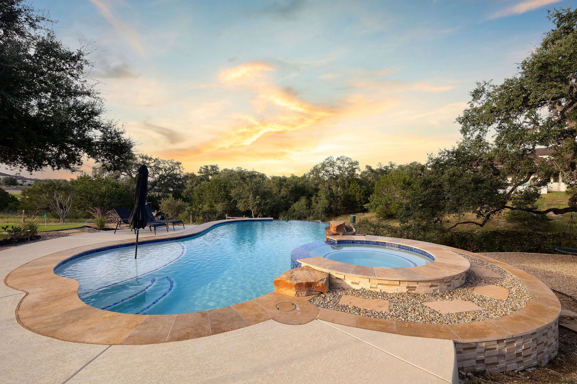 Backyard pool at sunset with a curved stone deck and surrounding trees